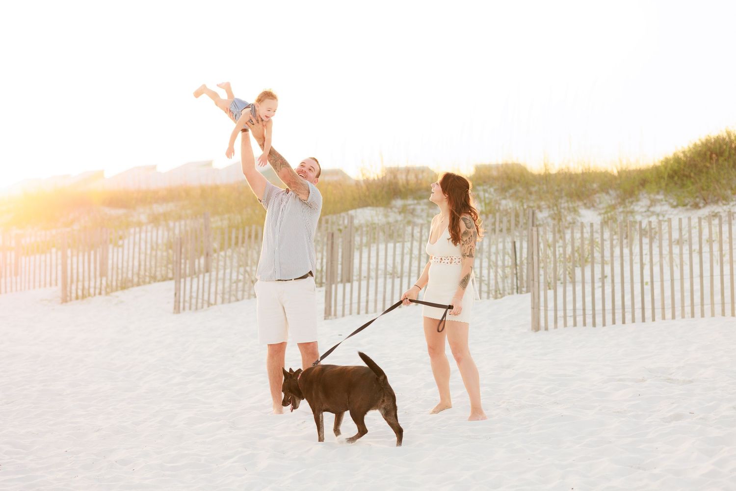 Father lifting baby in air while mother holds dog leash on sunny white sand beach.