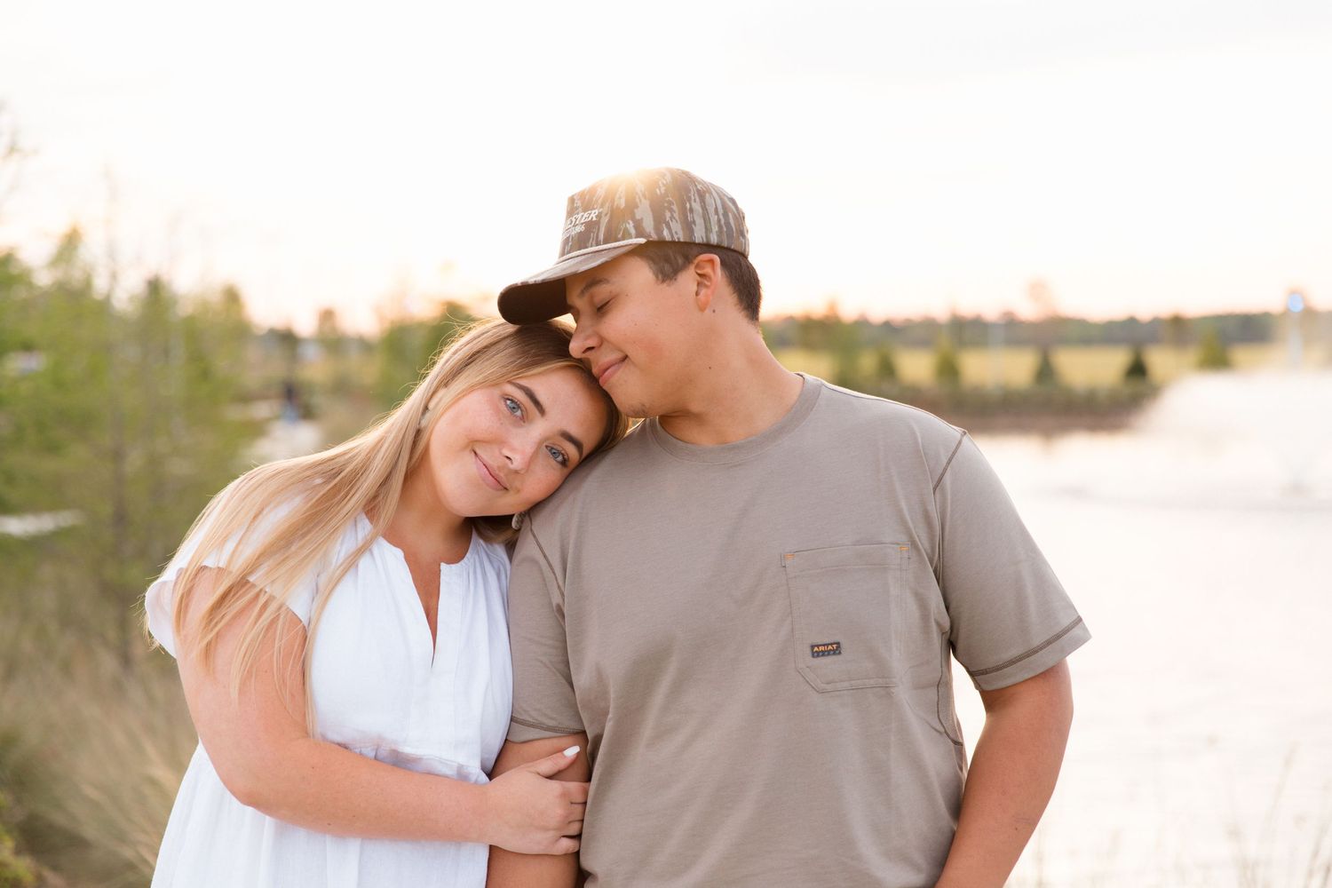 A young couple shares a tender moment outdoors, woman in white dress leaning on mans shoulder near a sunlit river.