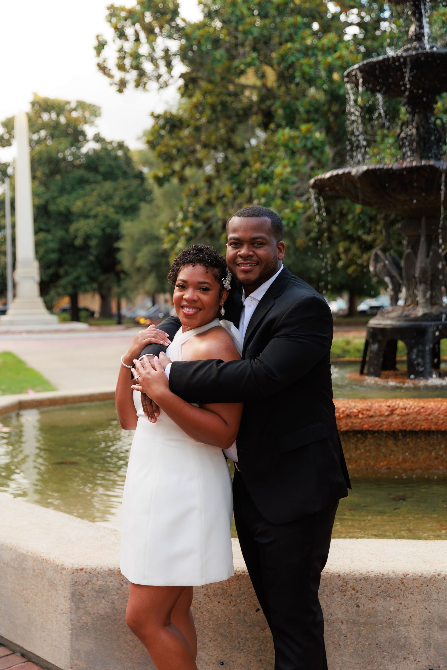 A couple poses lovingly by a tiered fountain, the woman in a white dress and the man in a black suit, in a park setting.