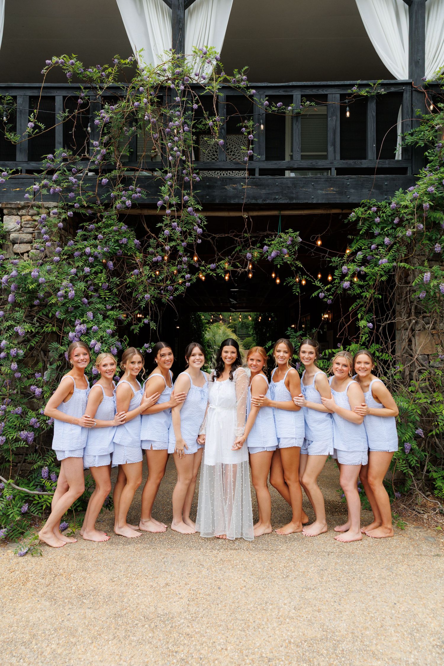 Bride in white robe poses with bridesmaids in matching blue pajamas outside a vine-covered venue.