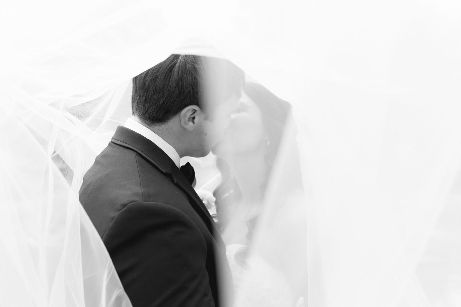 Black and white photo of groom leaning in to kiss bride beneath flowing wedding veil against bright background.