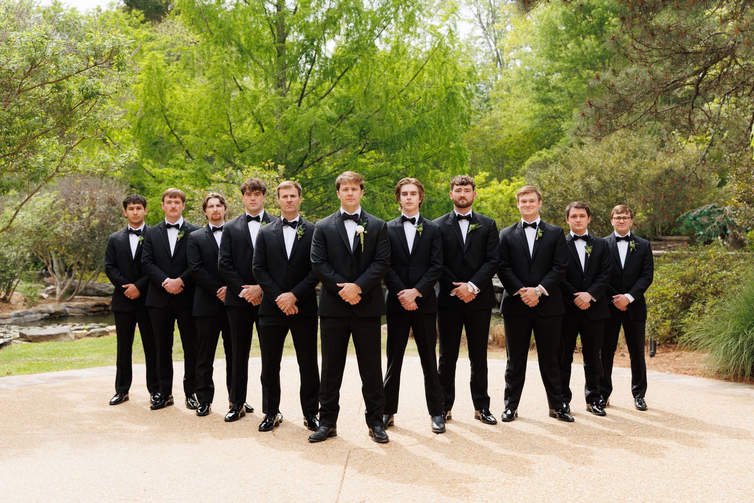 Group of ten groomsmen in matching black tuxedos and bow ties posing outdoors before a wedding ceremony.