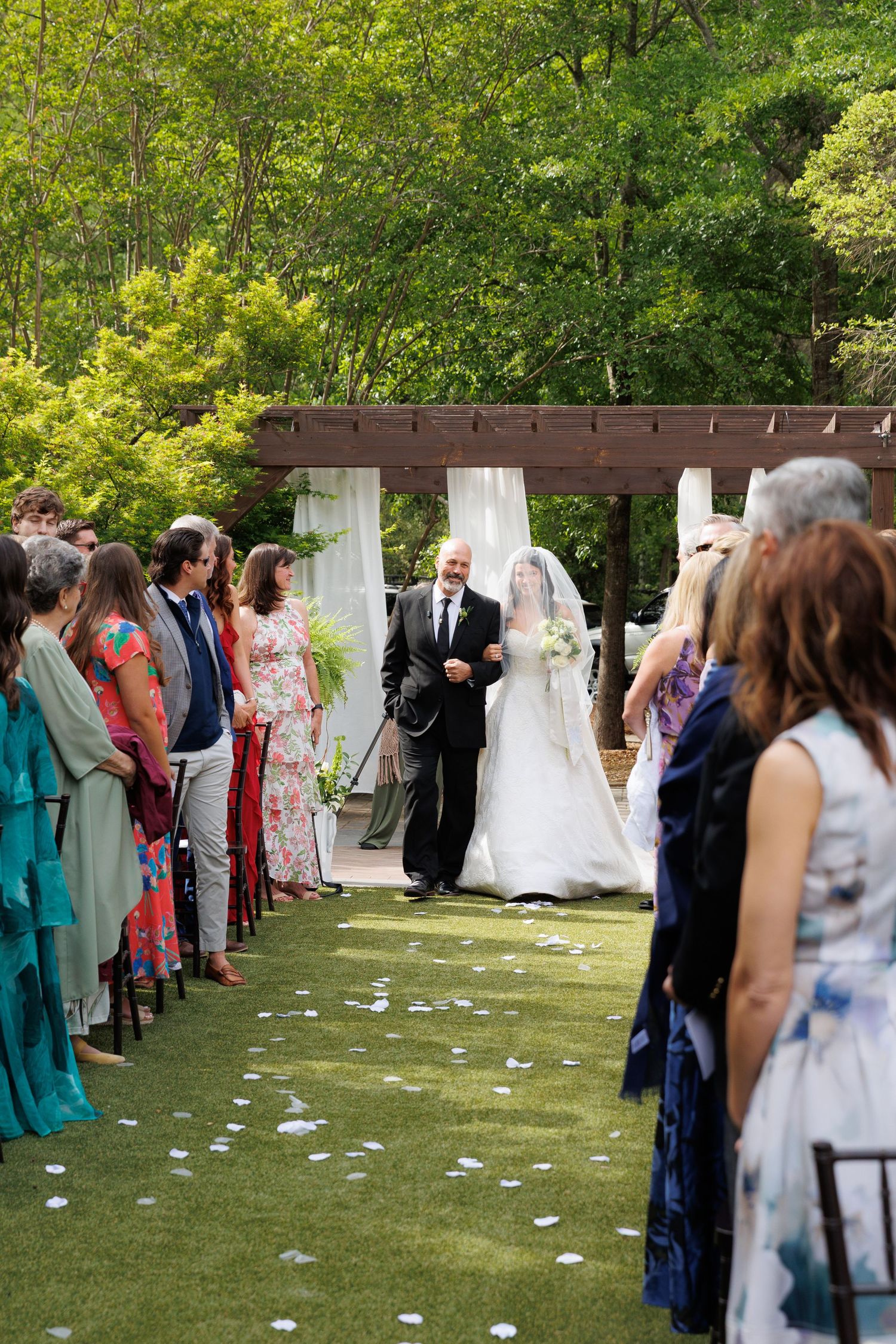 Bride walks down outdoor aisle with escort, guests standing, white petals on grass, pergola with draping in background.