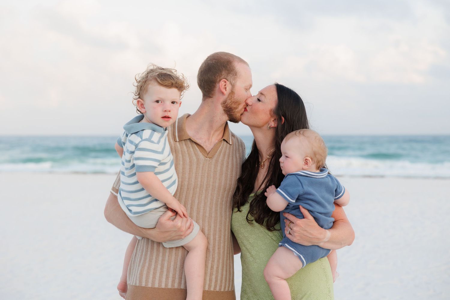 Parents kissing on white sand beach while each holding a young child with ocean waves behind.