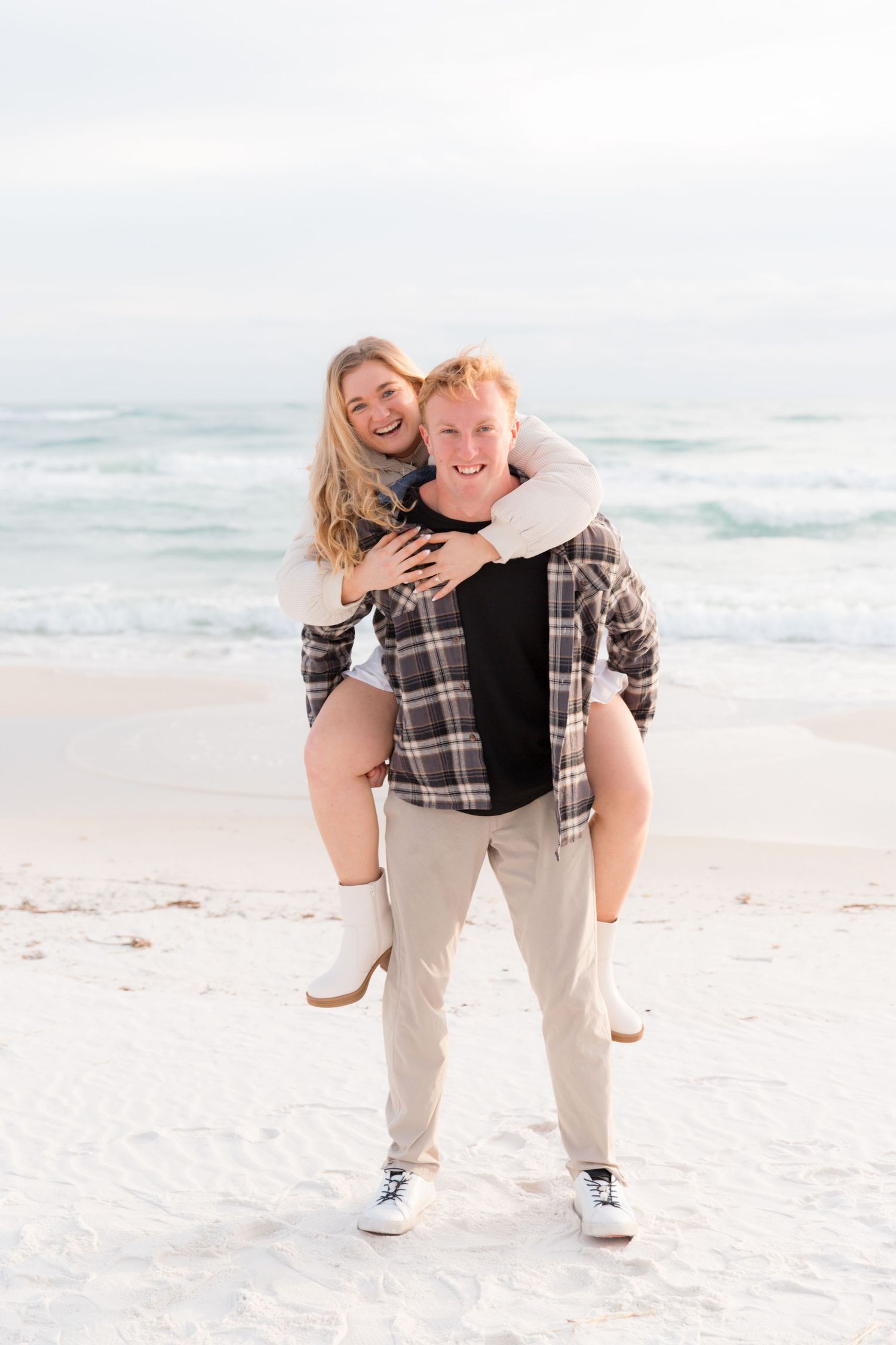 Playful couple having fun with piggyback ride on sandy beach with ocean waves behind them.