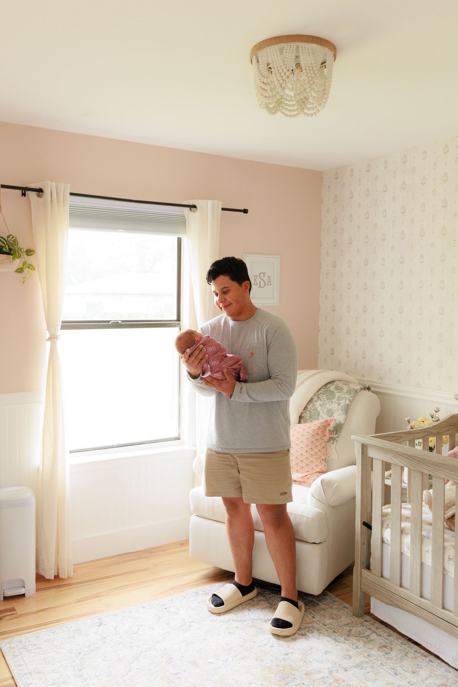 A person in gray sweater and shorts assembles nursery furniture in a bright room with white walls and floral wallpaper.