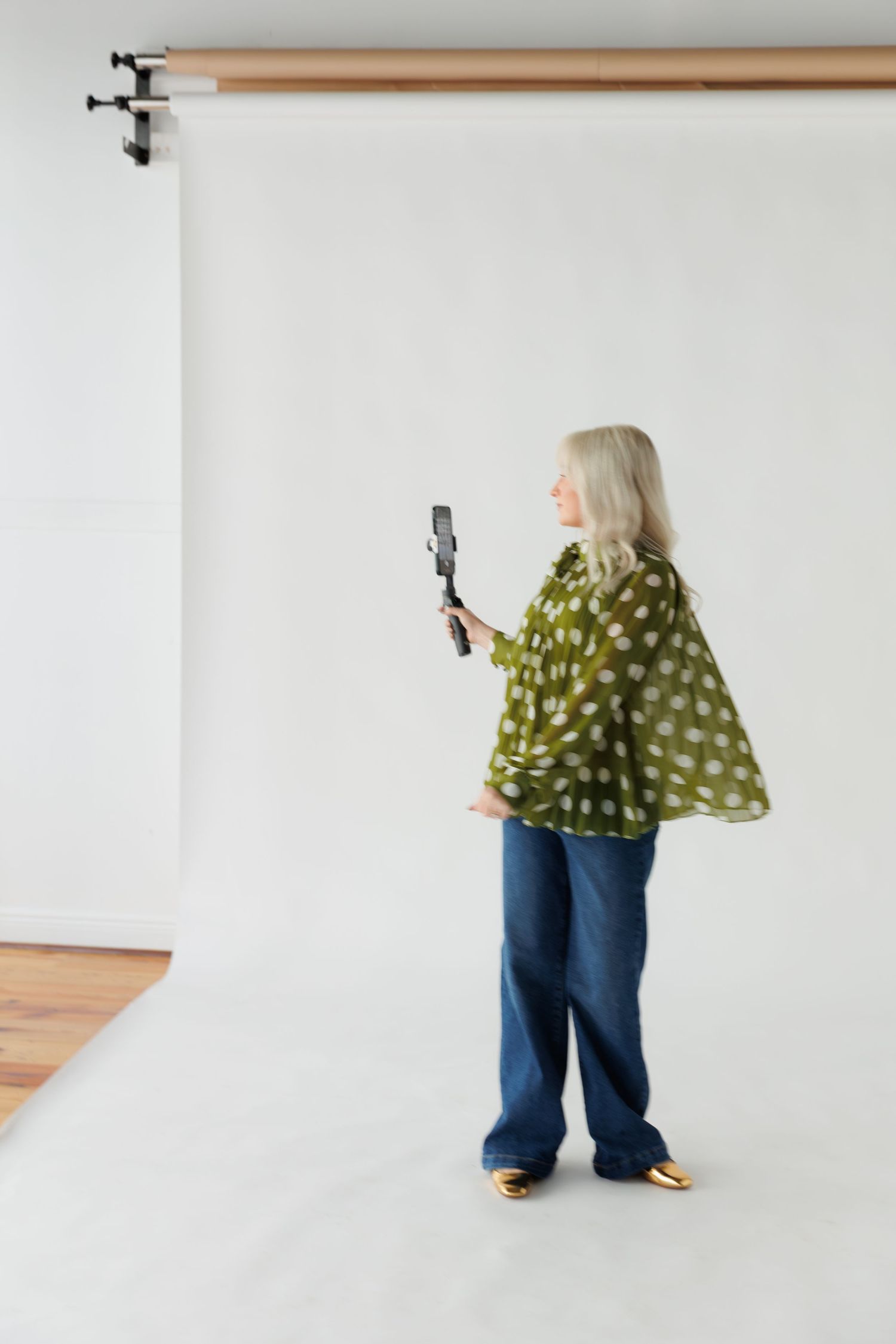 Person wearing a green polka dot top and jeans holds a camera while standing against a white backdrop in a photo studio.