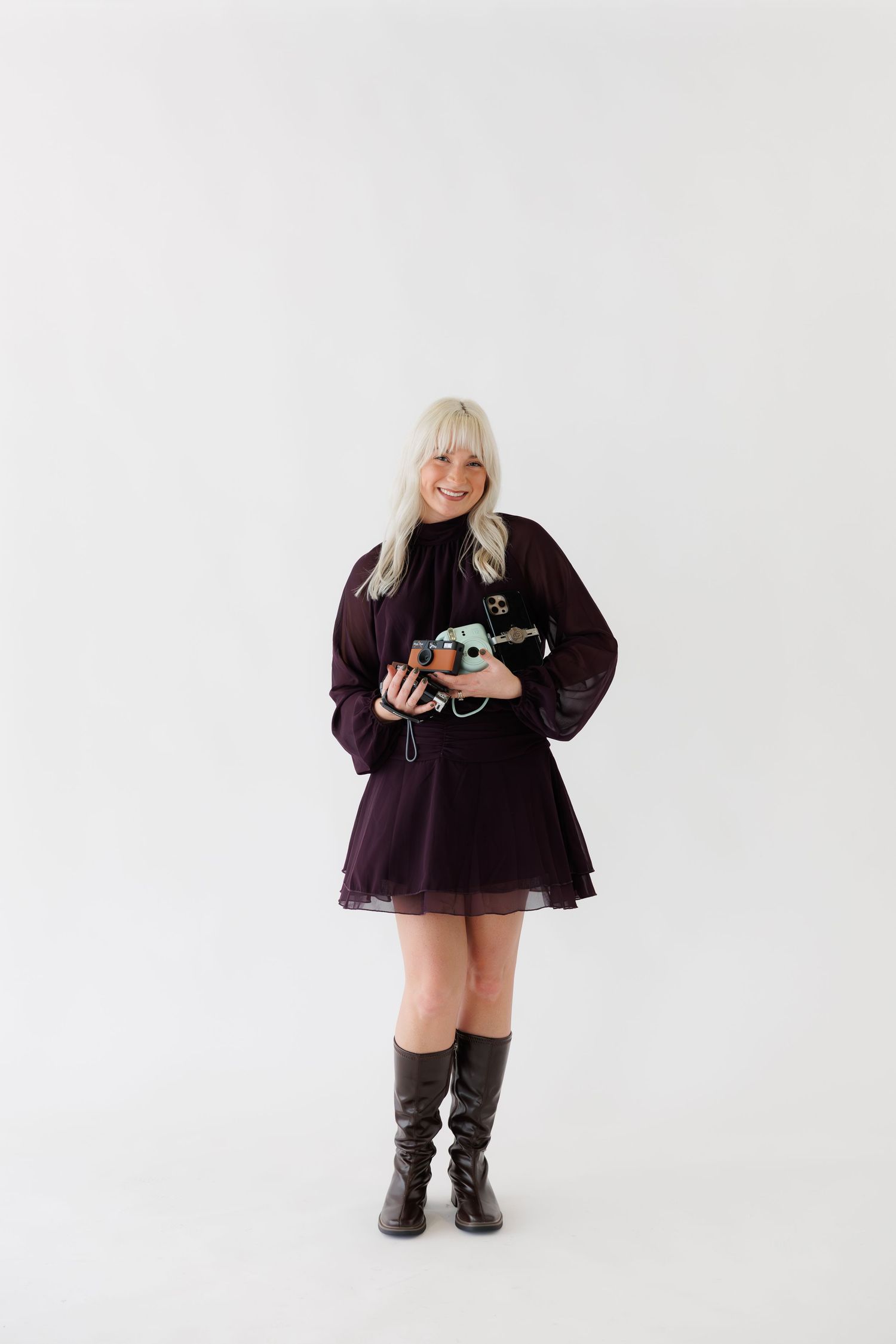A person in a purple dress holds a camera while standing against a white studio backdrop.