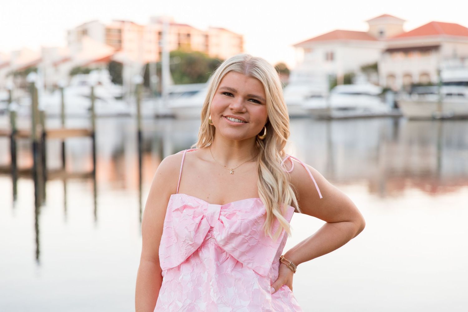 Woman in pink floral dress poses outdoors near marina with boats and buildings in background.