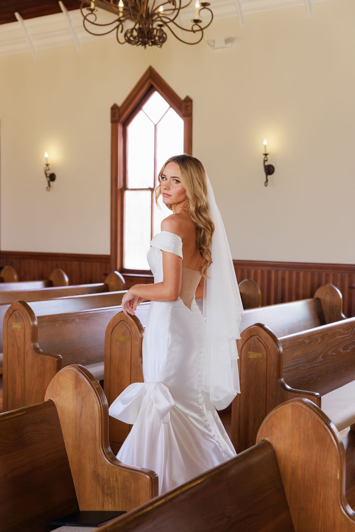 A bride in a classic white gown creates artistic poses among traditional wooden church pews.