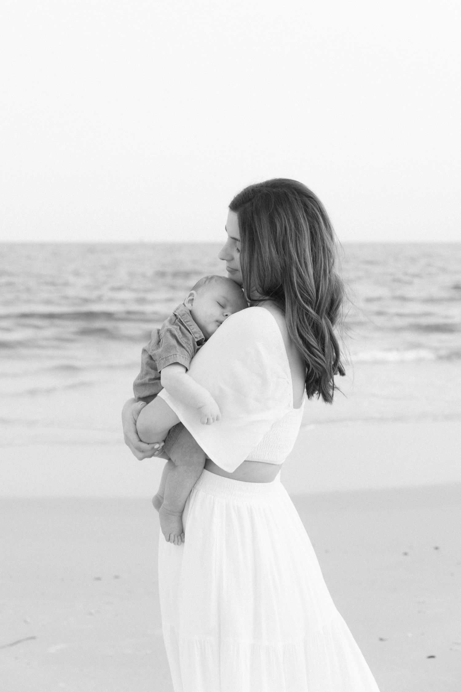 Black and white photo of mother in white dress cradling newborn baby on beach.