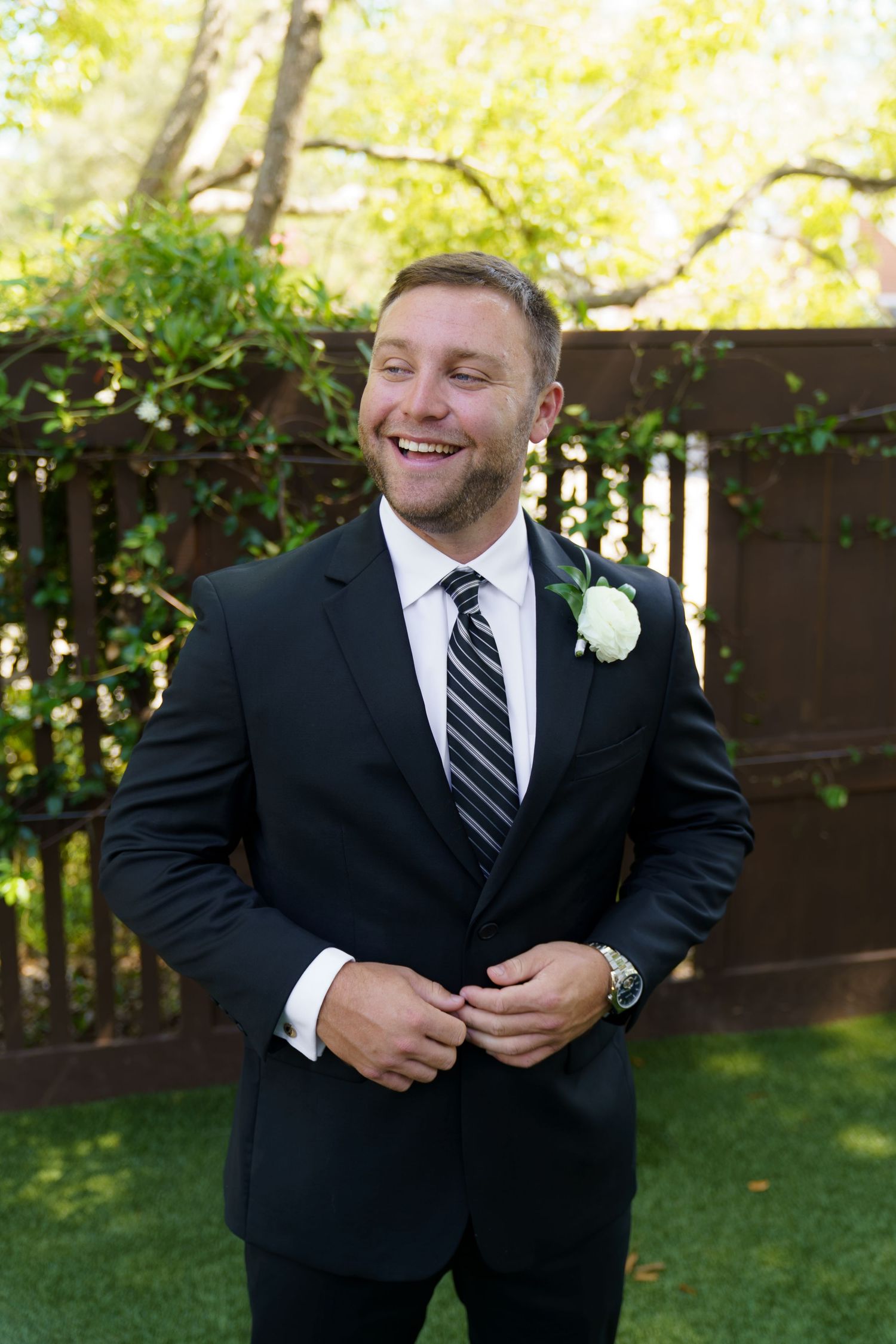 Smiling groom in black suit with white boutonniere buttoning jacket outdoors at wedding ceremony.