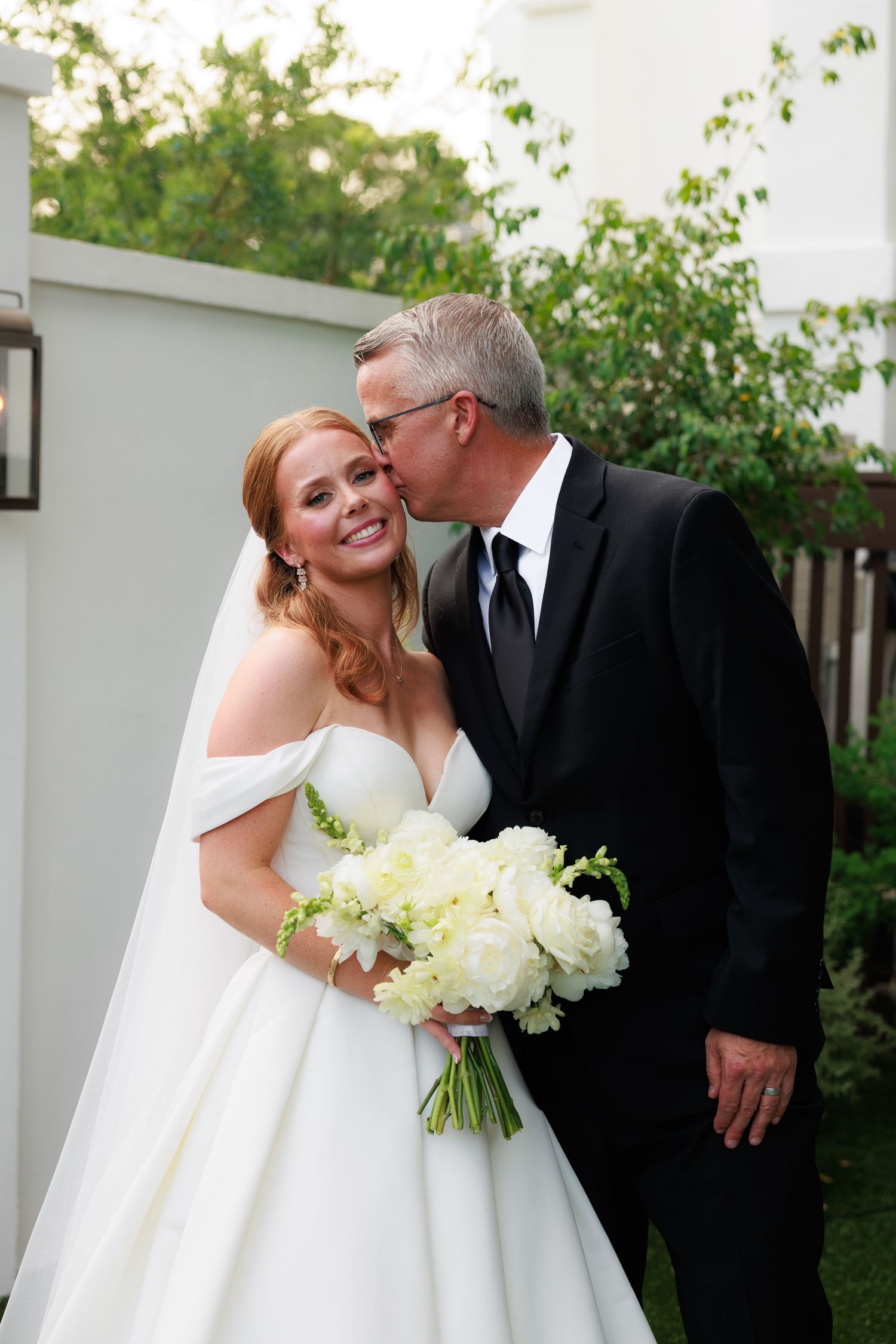 Bride in off-shoulder white gown holding bouquet receives kiss from groom in black suit outdoors.