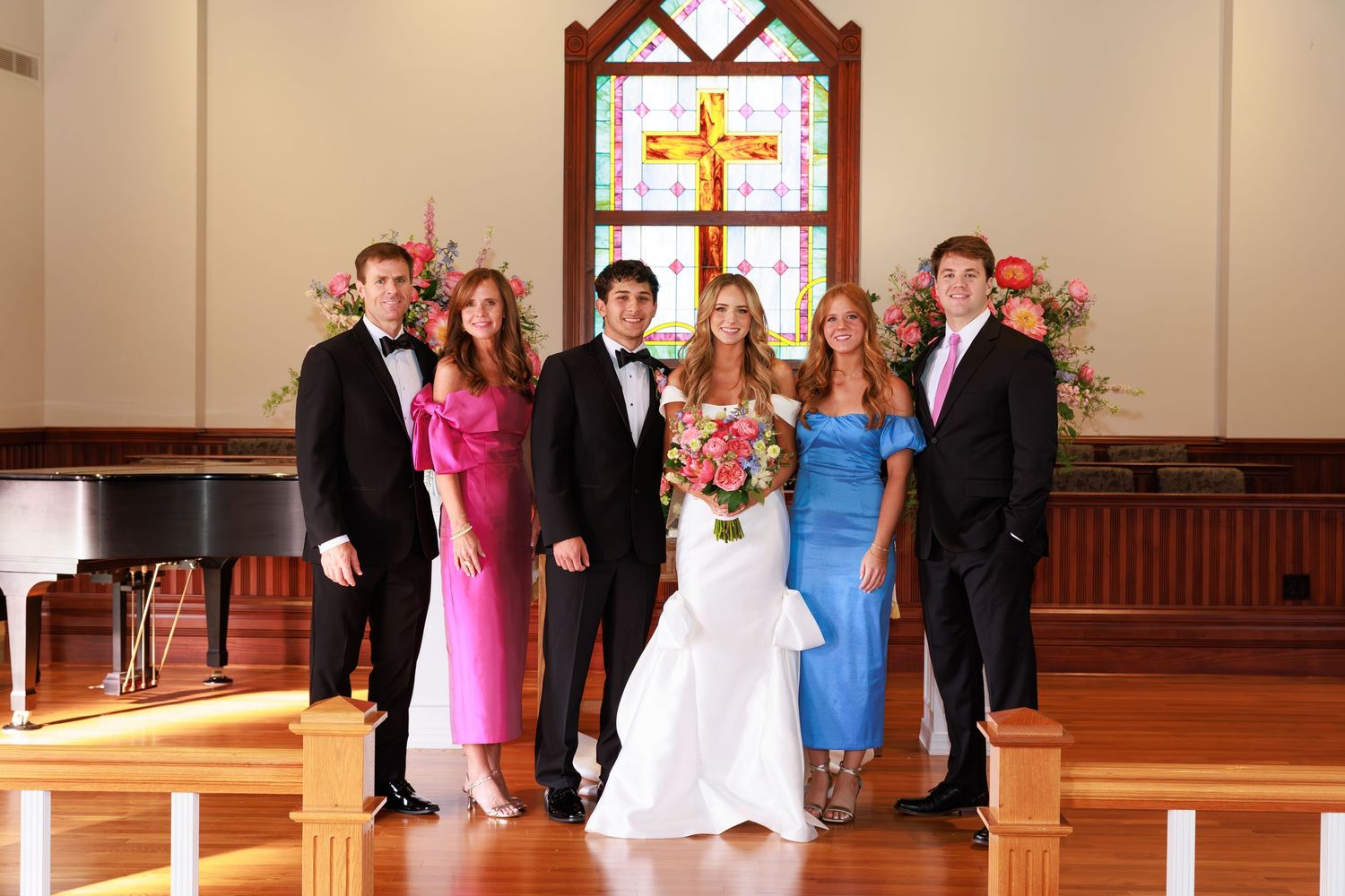 Small wedding party gathering in front of stained glass cross window in church sanctuary.