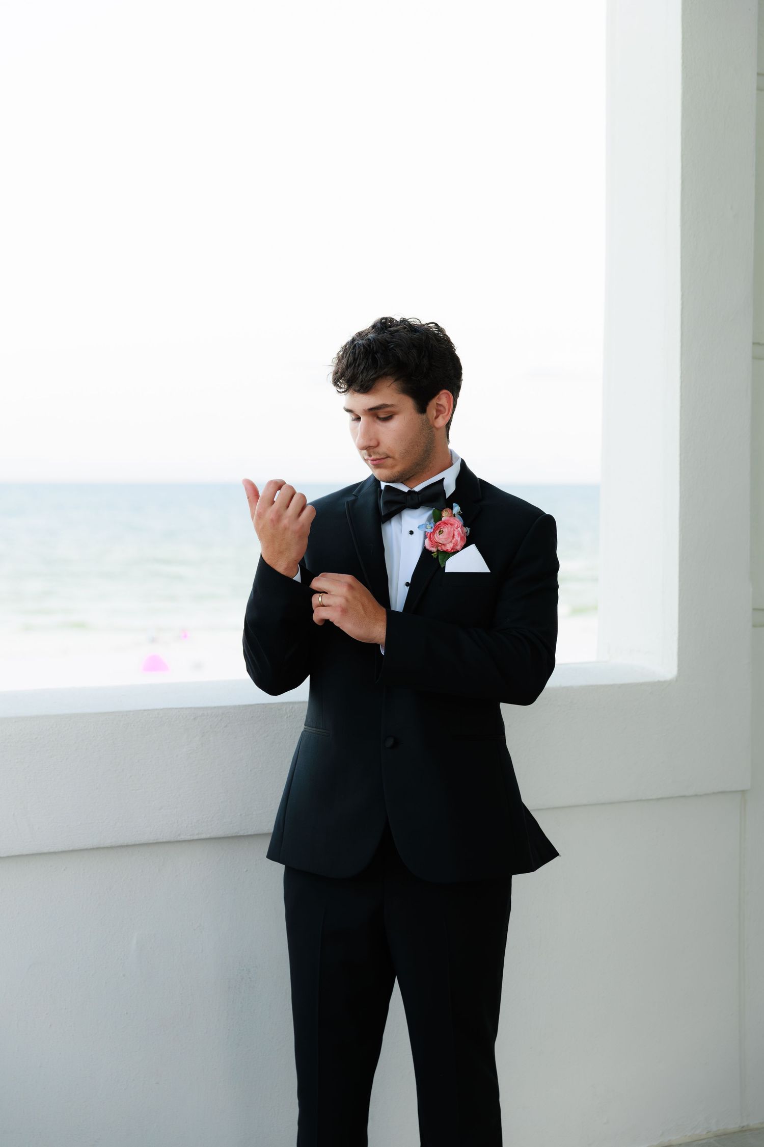 A groom in a black tuxedo adjusts his bowtie while standing in a bright white coastal venue.