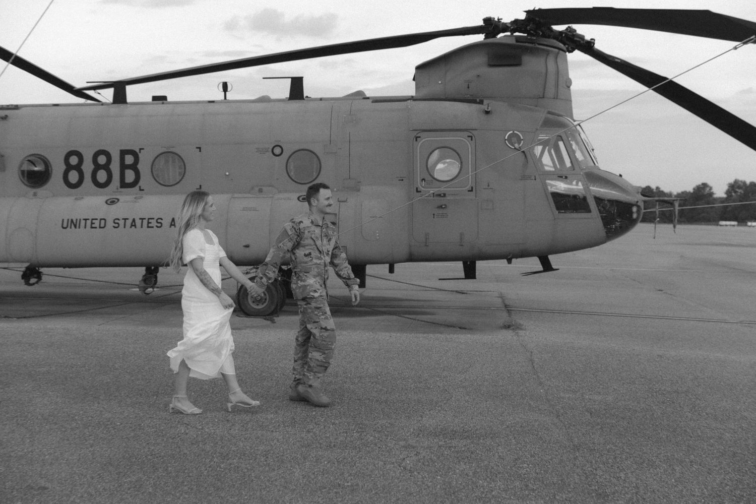 Black and white photo of a couple walking near a military helicopter.
