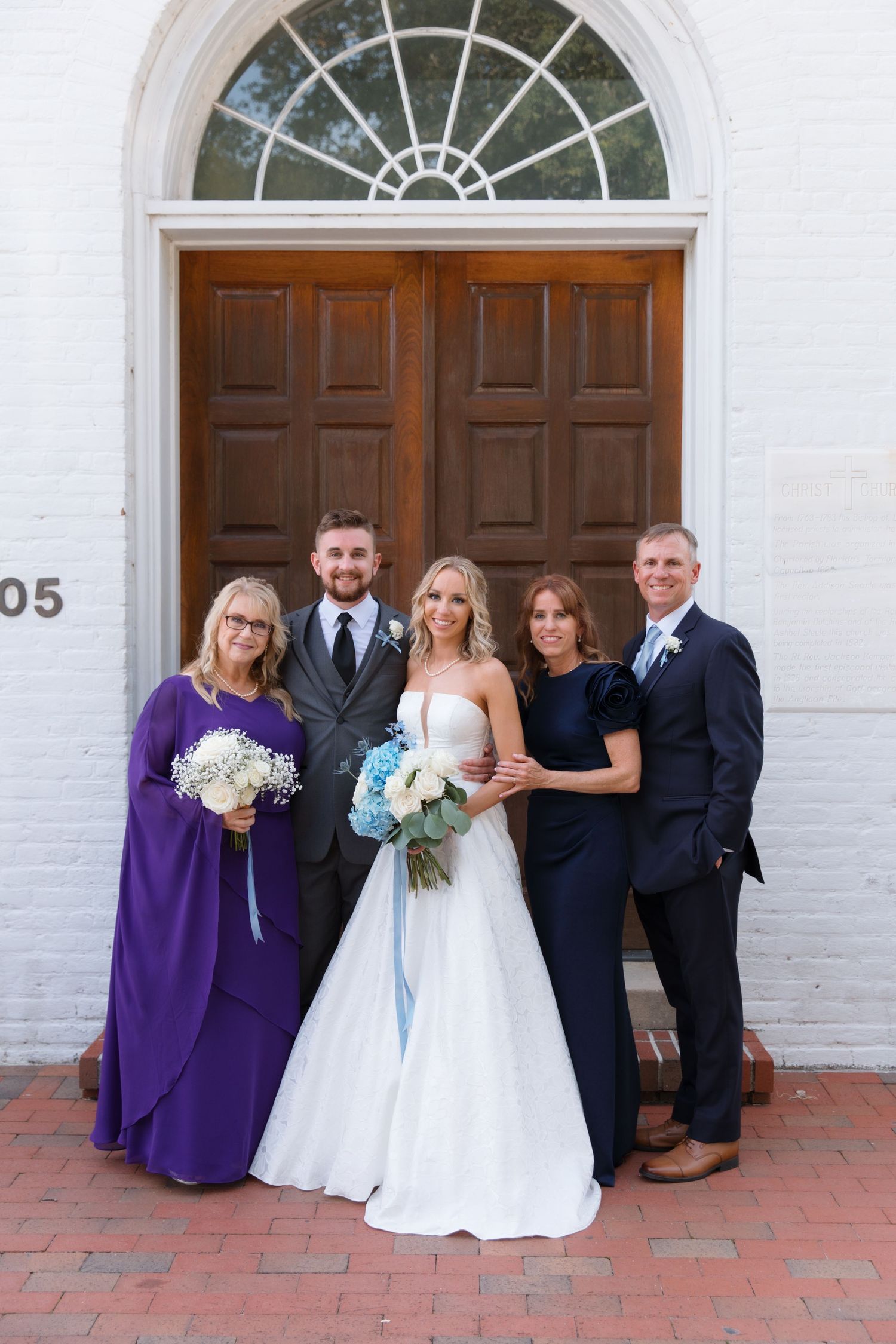 Wedding party of five people posed formally in front of white church with classic wooden doors.