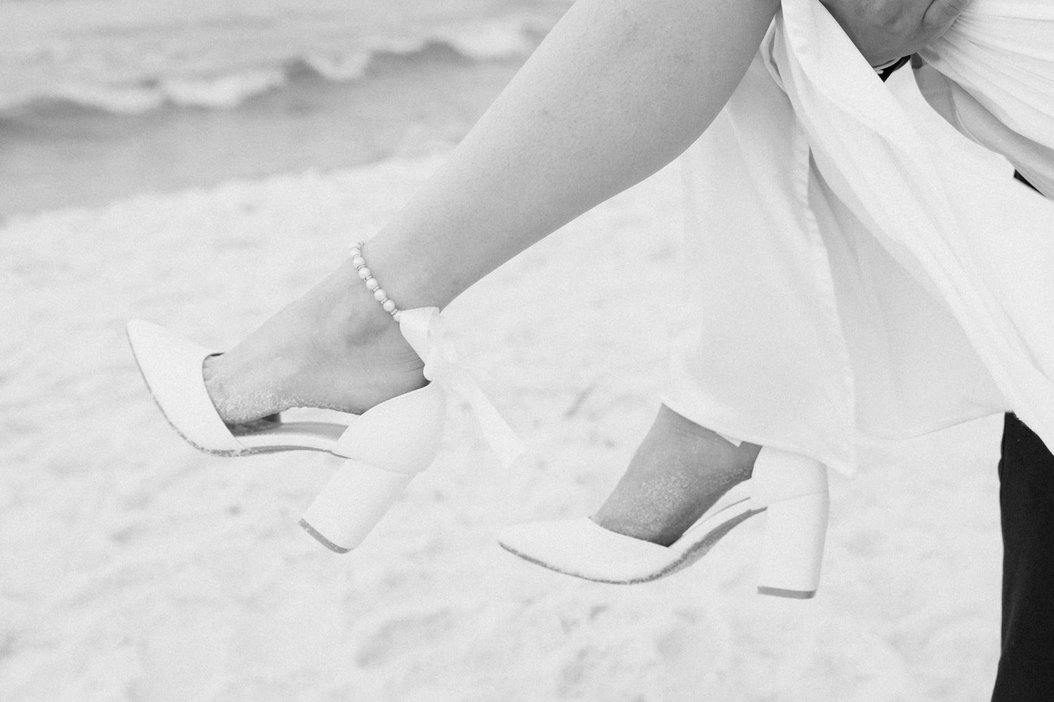 Close-up of bride's feet wearing white block heel sandals on beach sand.