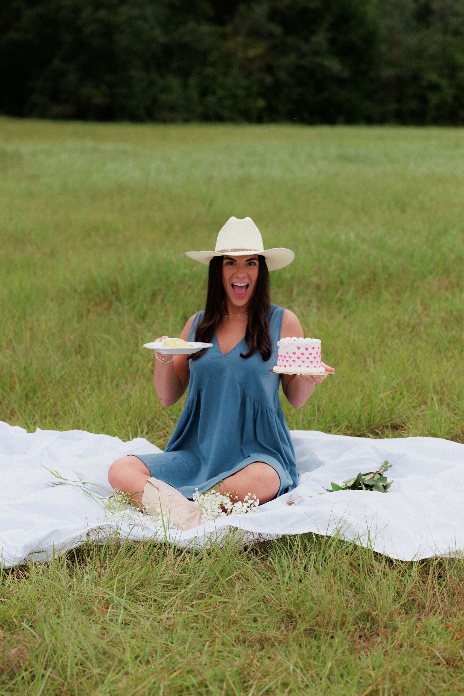 Person in western attire having a picnic with cake on a white blanket in a grassy field.