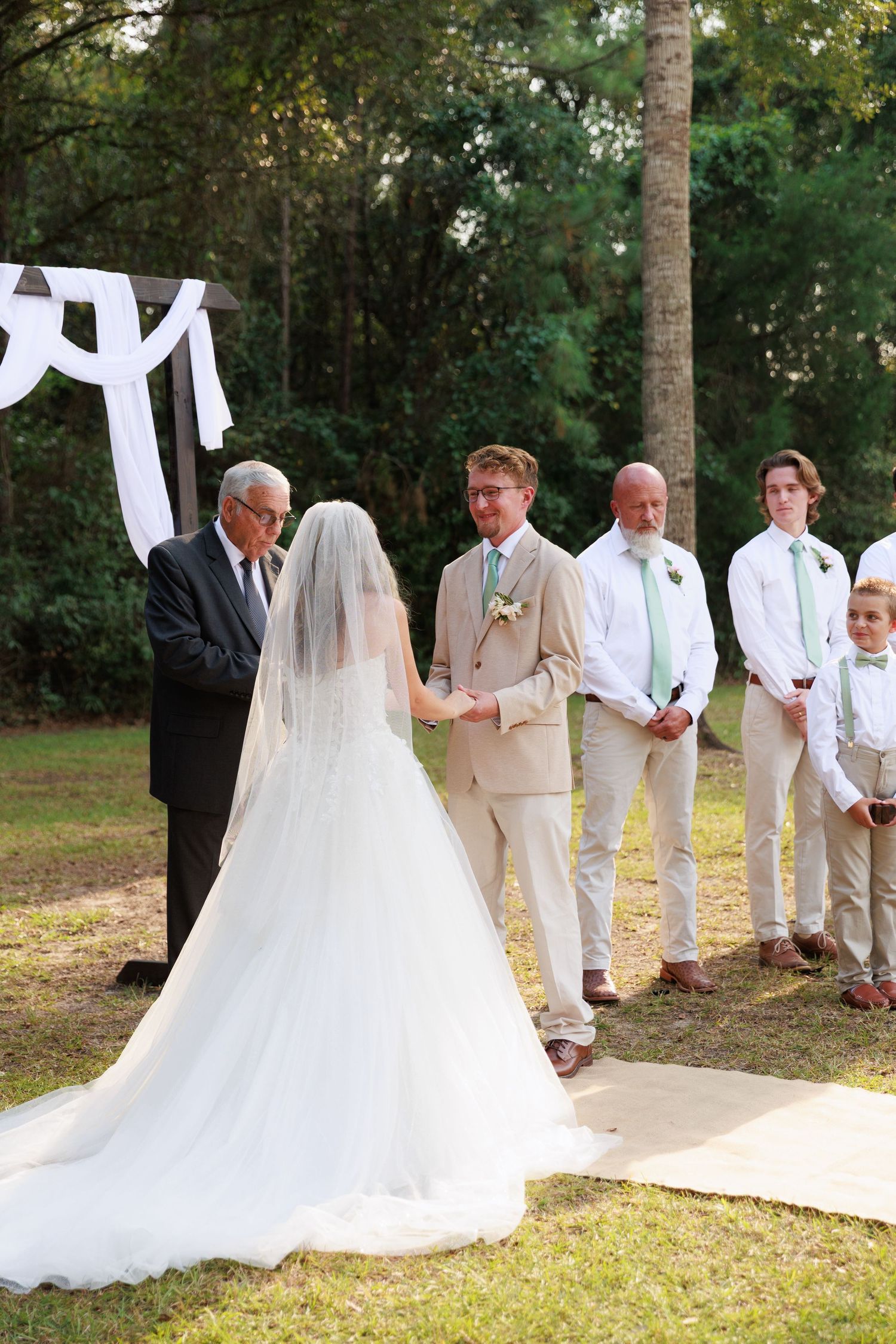 A wedding ceremony taking place beneath a white draped arch with attendants in light colored attire.