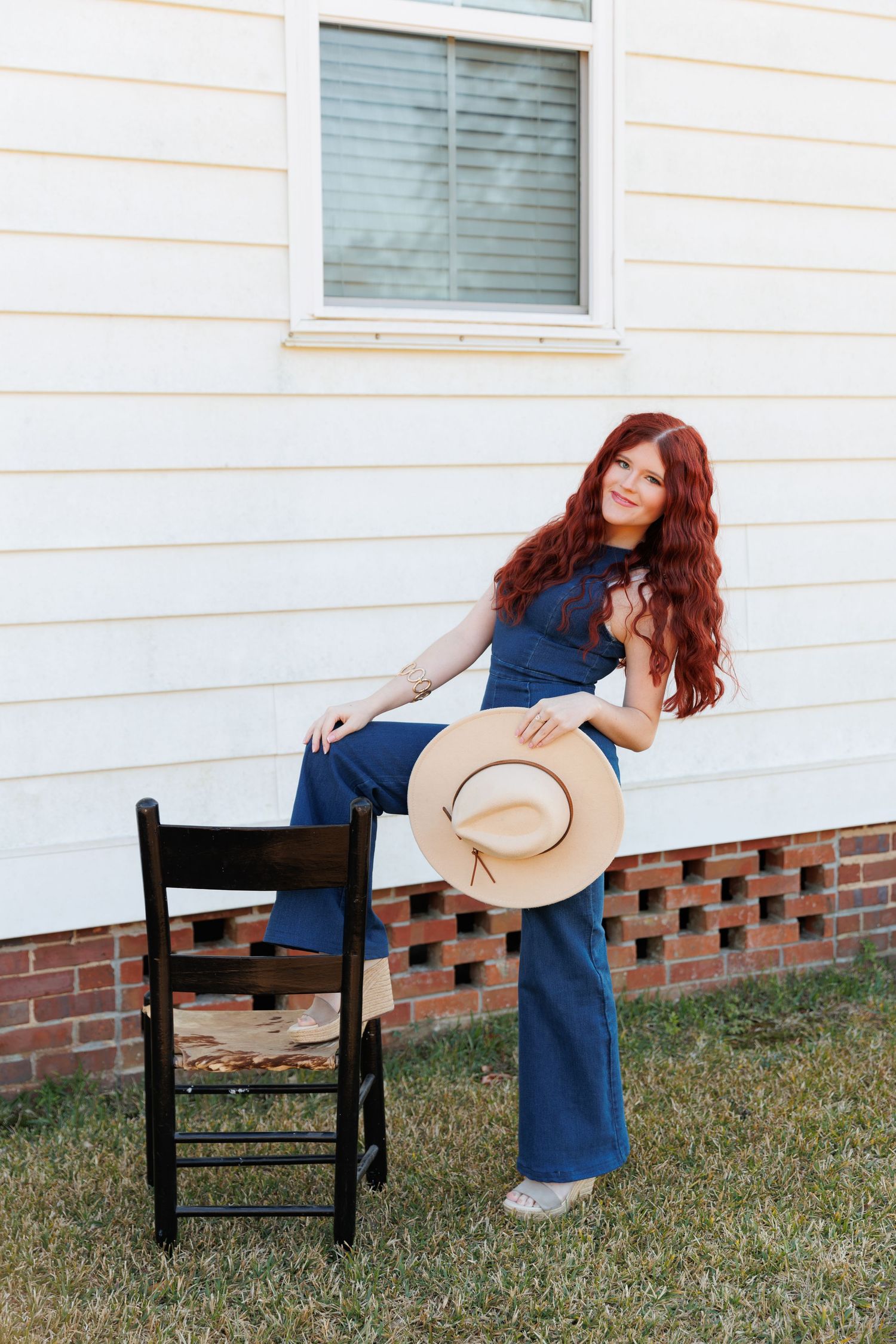 A person in a blue dress sits on a black wooden chair outdoors against a white house with brick foundation.