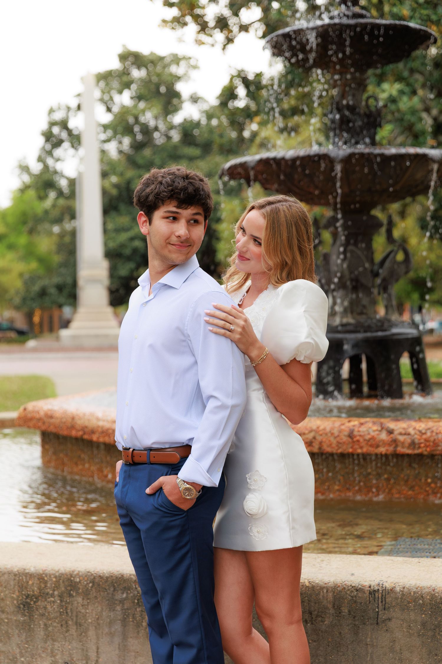 A couple shares a tender moment by a fountain in a scenic park setting.