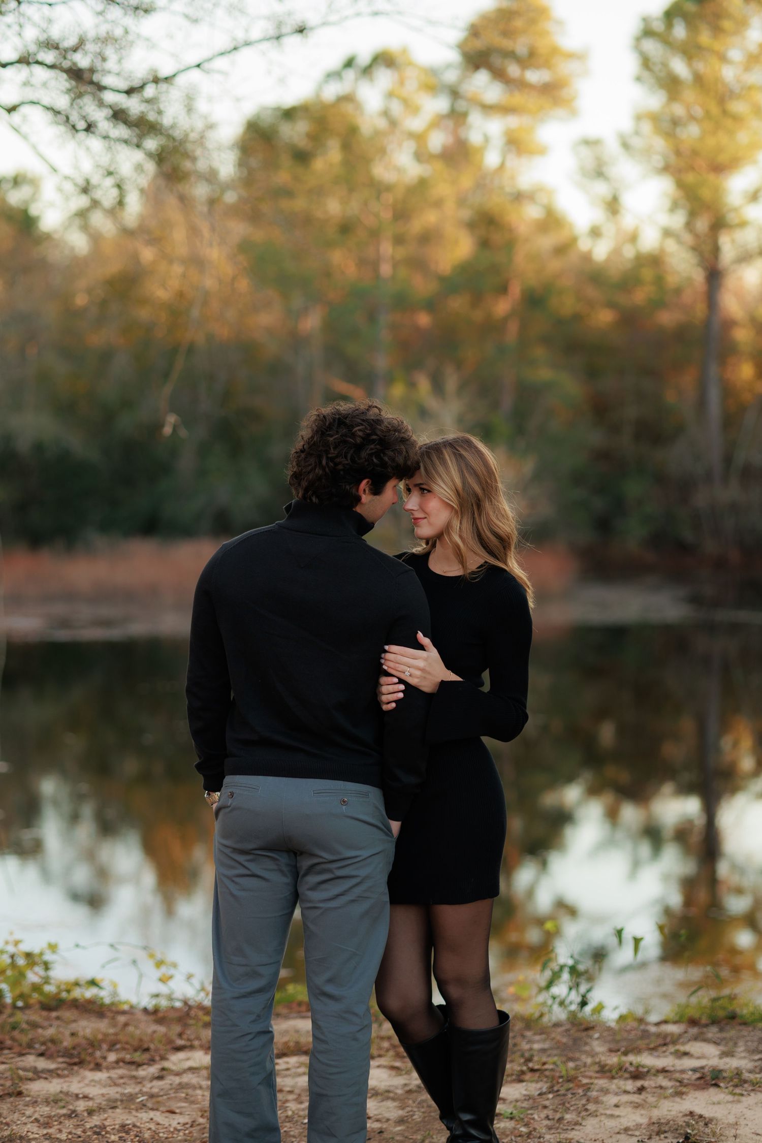 Couple in matching black outfits embracing near a tranquil lake surrounded by autumn trees.