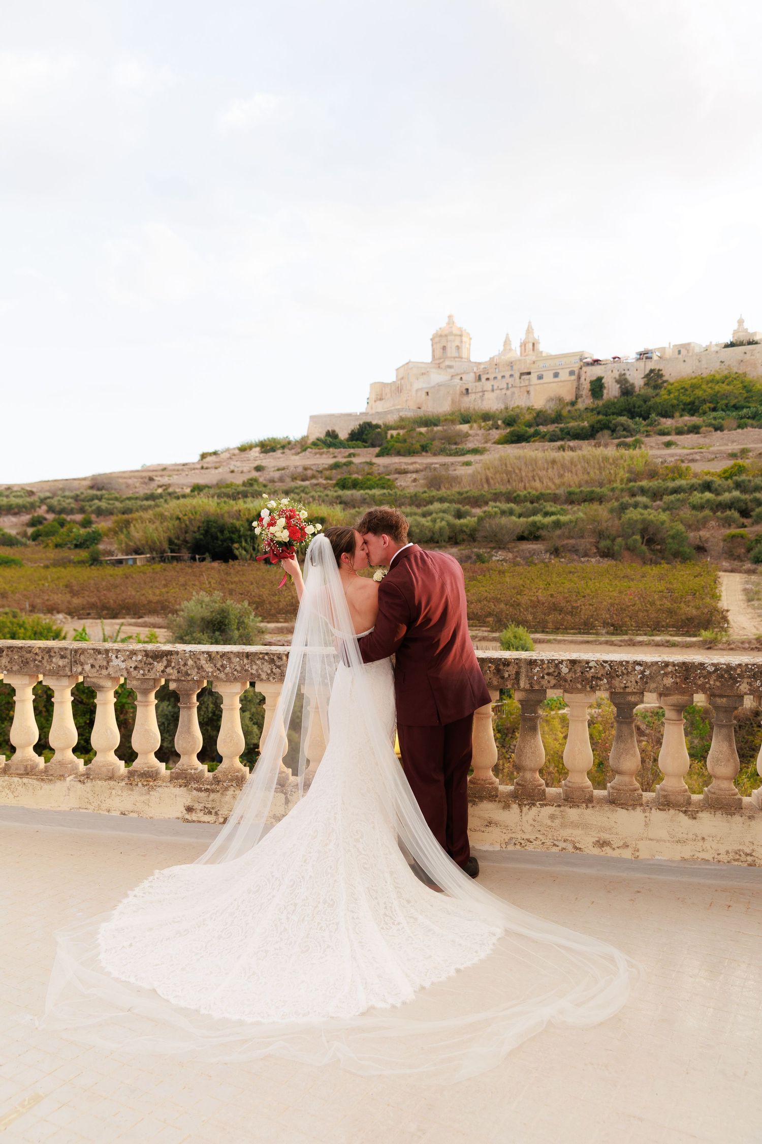 A romantic wedding couple embraces on a stone balcony overlooking scenic countryside views in Malta.