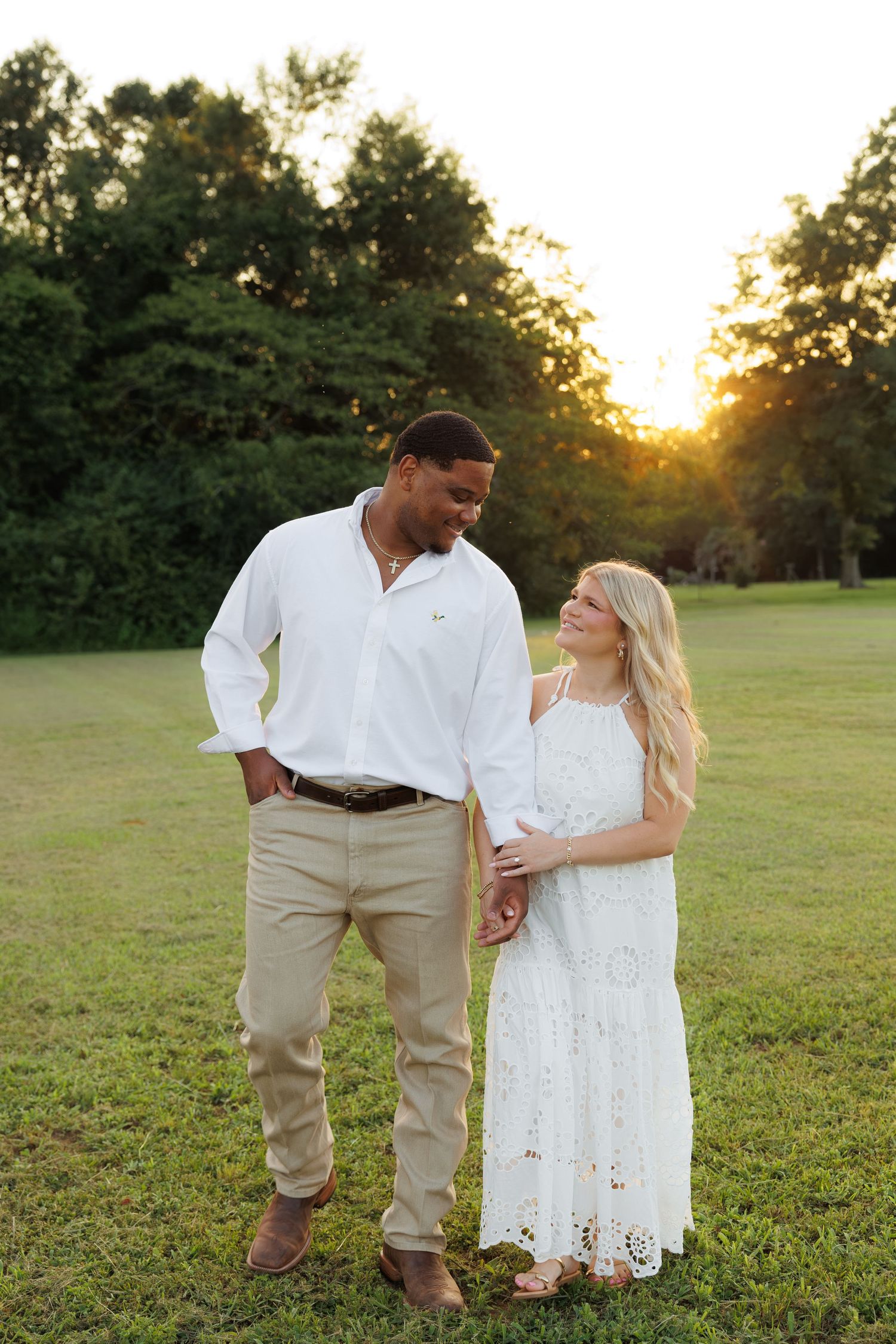 Couple in white outfits holding hands at golden hour sunset in a green field with trees in background.