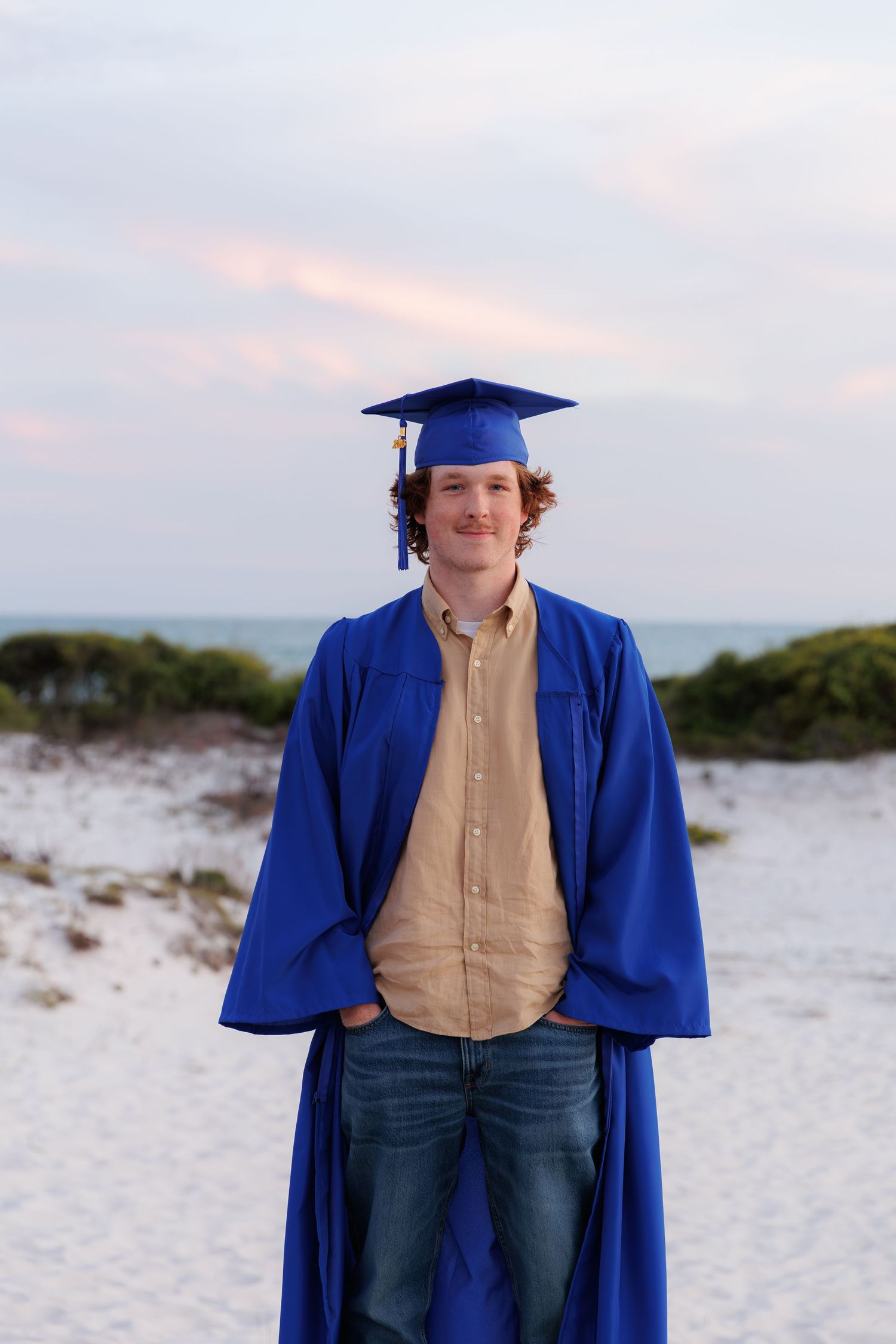 Smiling graduate in blue cap and gown standing on sandy beach at dusk.