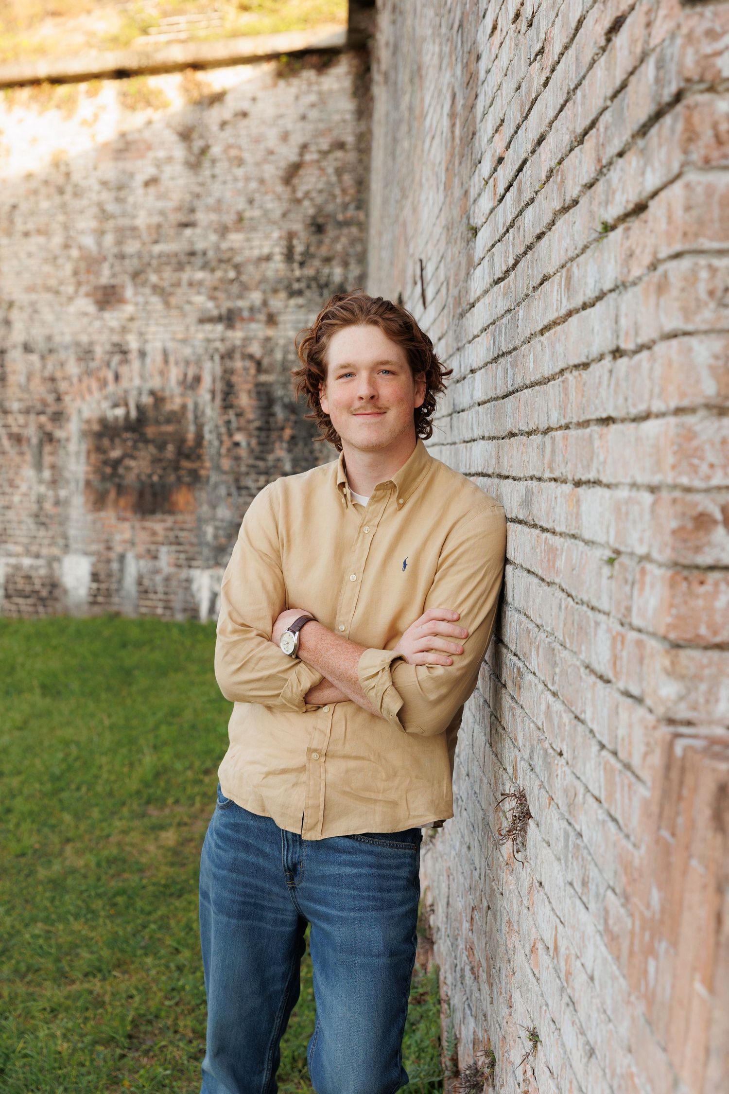 Young man in yellow shirt with arms crossed leaning against brick wall outdoors.