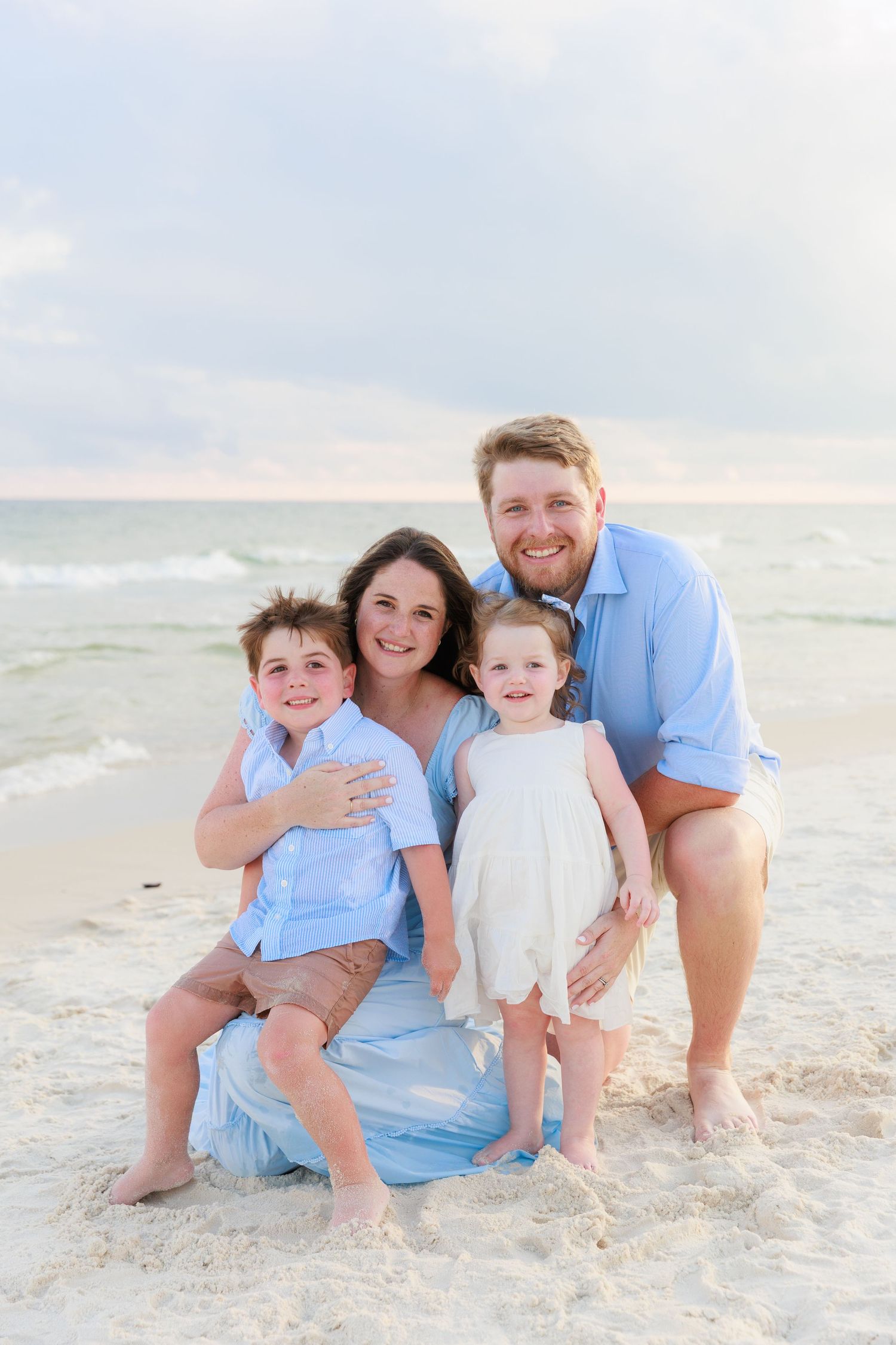 A smiling family of four poses together on a sandy beach at sunset.