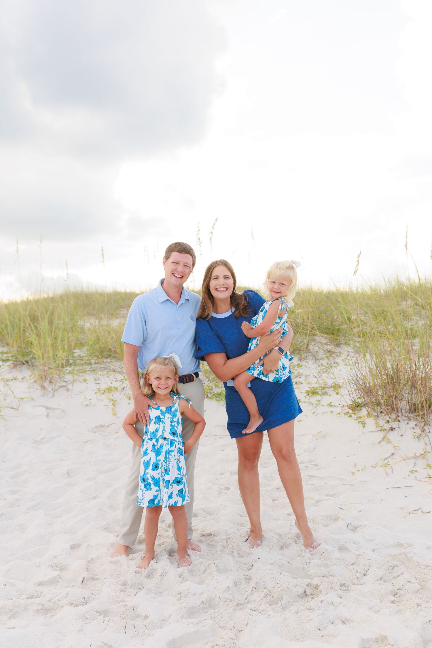 Family of four posing on sandy beach dunes, parents and two young girls wearing coordinating blue outfits.