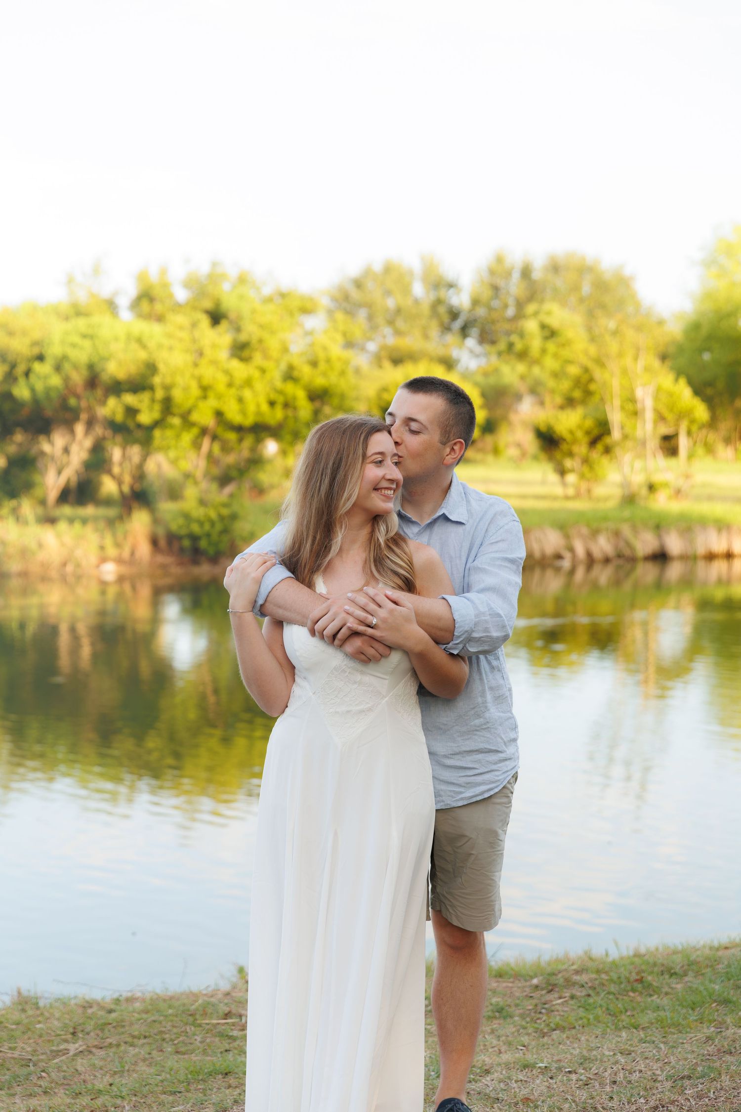 Couple embracing by a serene pond, man kissing woman's cheek, she wears white dress, lush green trees in background.