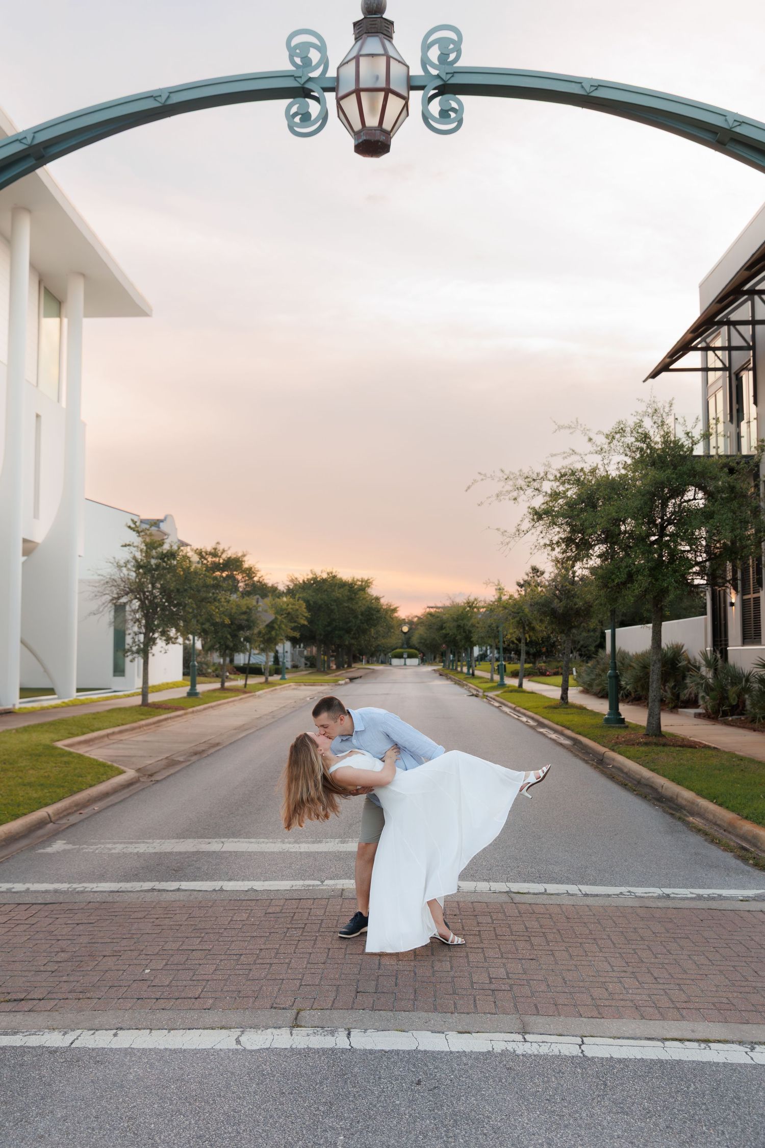 Couple sharing a romantic dip kiss under an arch streetlamp at sunset on a tree-lined boulevard.