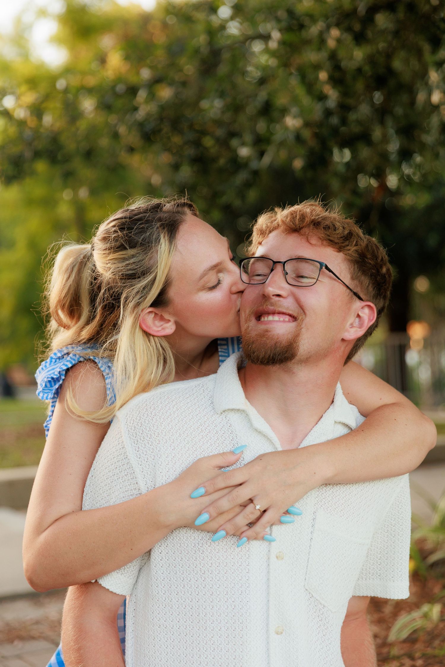 A smiling woman in a blue gingham dress hugs a laughing man from behind during an outdoor engagement photo session.