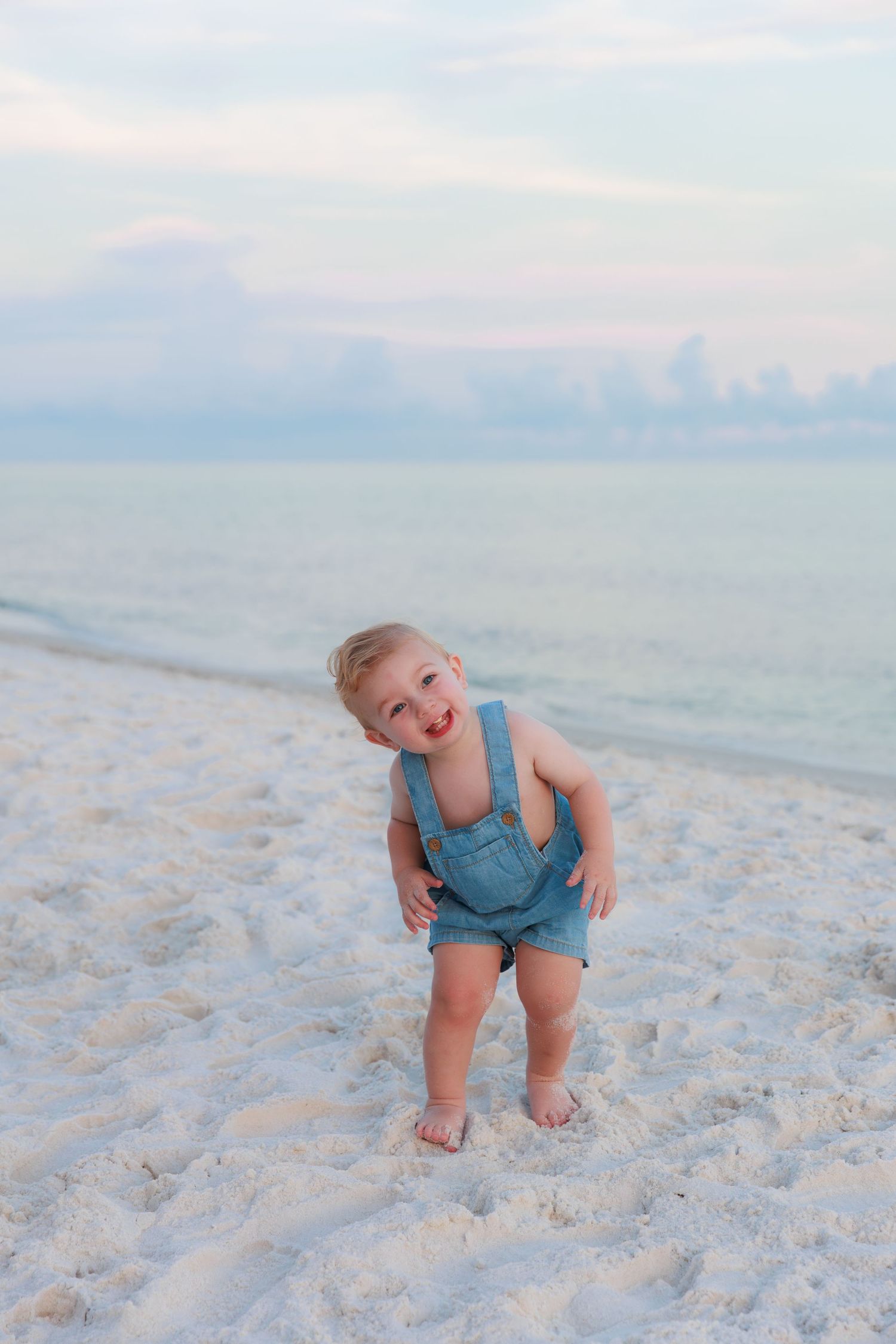 Toddler in denim overalls standing on white sand beach with calm turquoise water in the background.