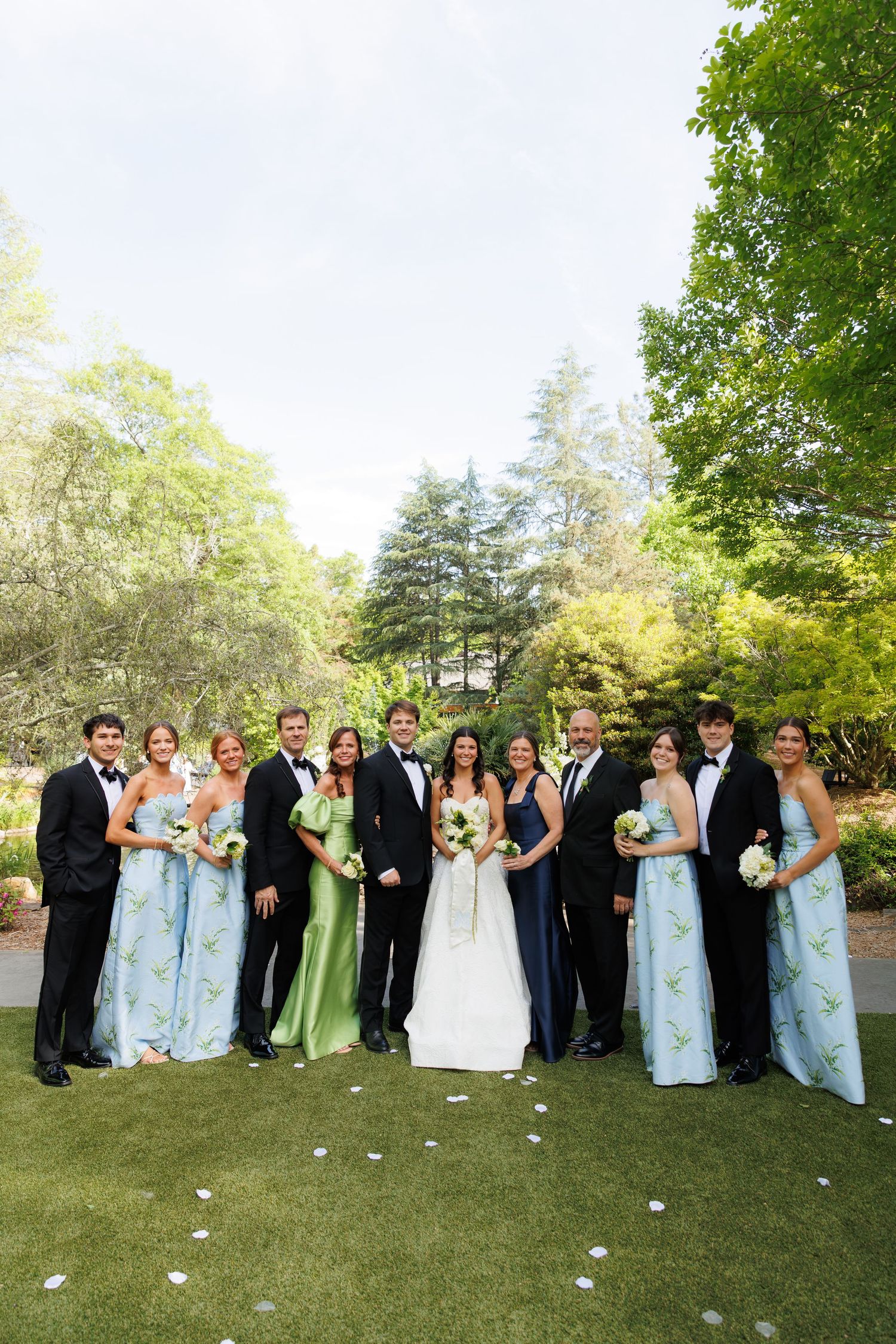 Wedding party poses outdoors in lush garden, bridesmaids in blue floral and green gowns, groomsmen in black tuxedos.