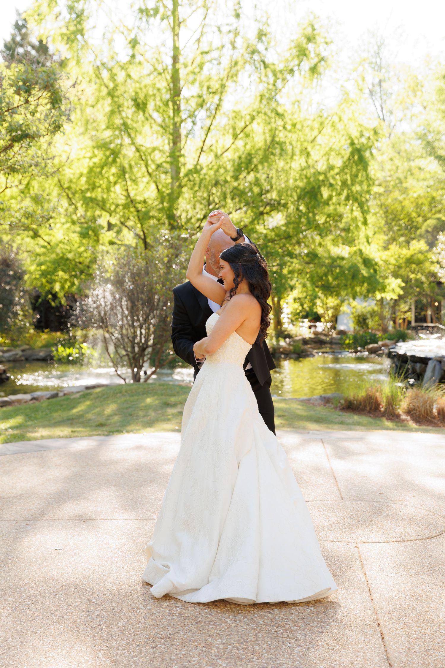 Groom twirls bride in elegant white gown during an outdoor dance near a tranquil garden pond.