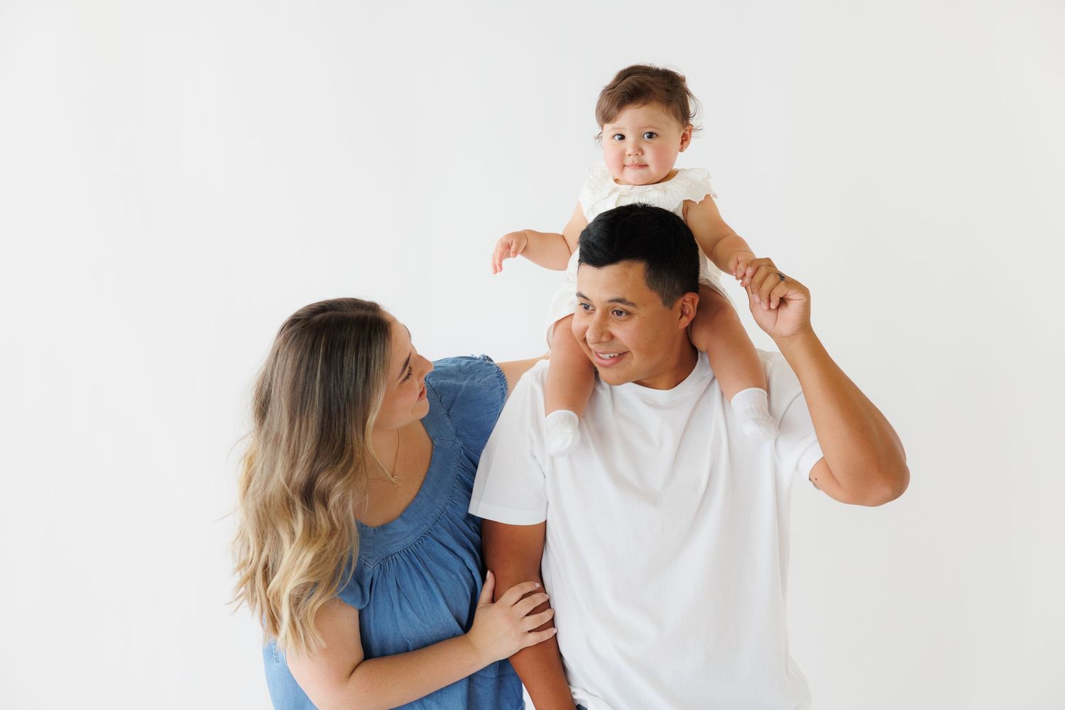 Happy family photo with mom, dad, and baby on shoulders smiling against white background.