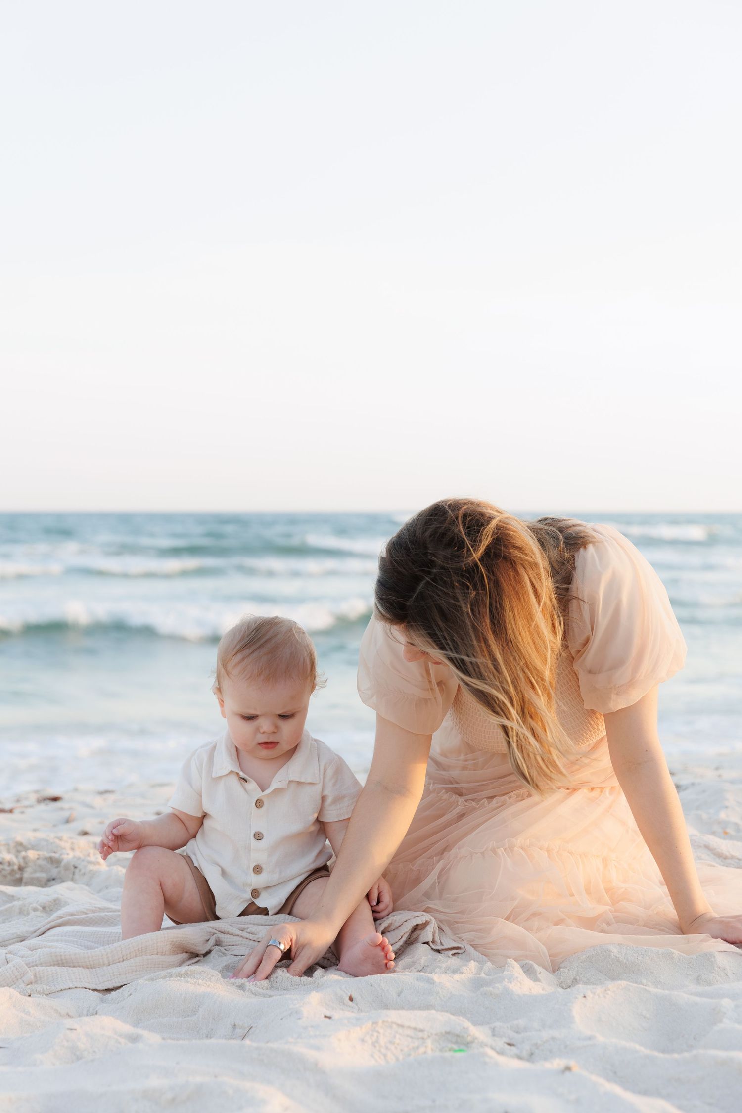 Mother in pink dress playing with baby on white sand beach by ocean.