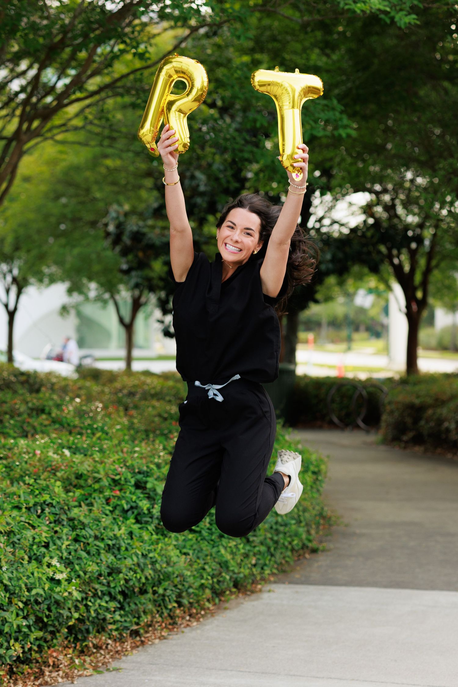 Woman in black scrubs jumping joyfully outdoors while holding gold balloon letters RT, celebrating becoming a respiratory therapist.