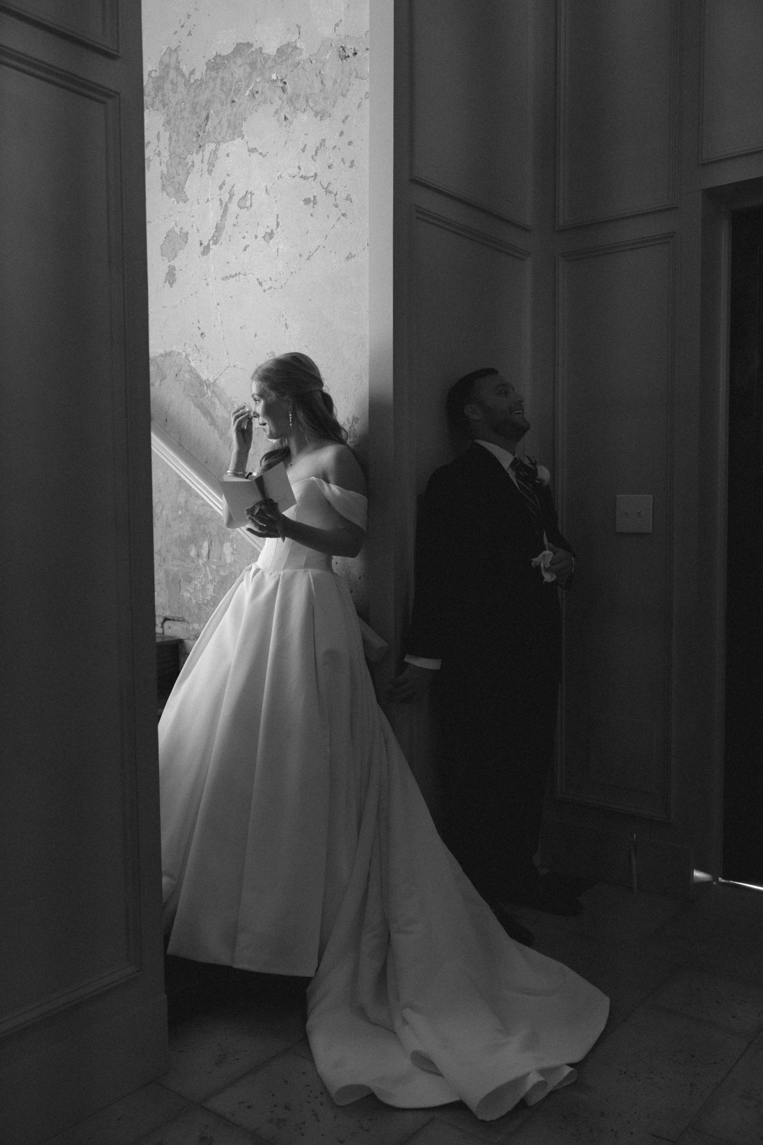 Black and white photo of bride in off-shoulder gown and groom in suit standing back-to-back before wedding ceremony.