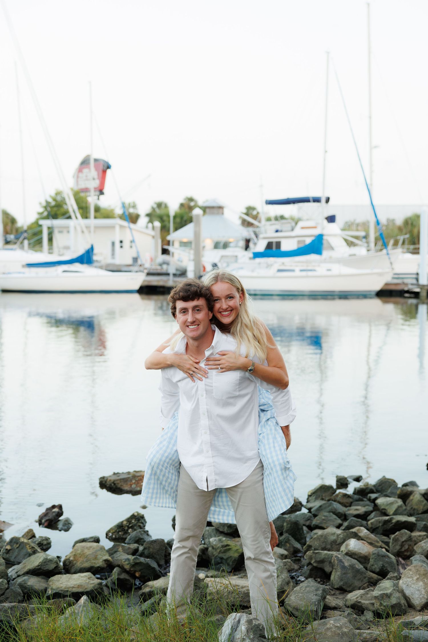 A smiling couple poses together on rocky shoreline with sailboats and marina reflecting in calm water behind them.