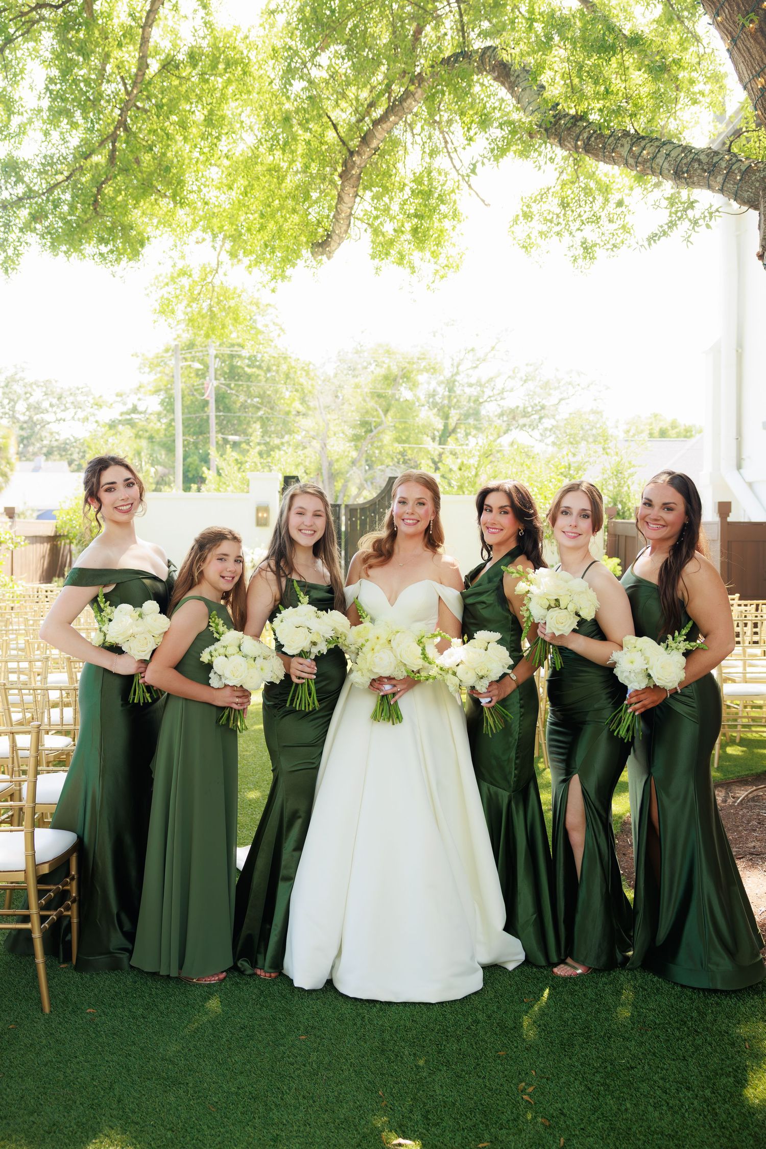 Bride in white gown poses with six bridesmaids in emerald green dresses holding white bouquets outdoors.