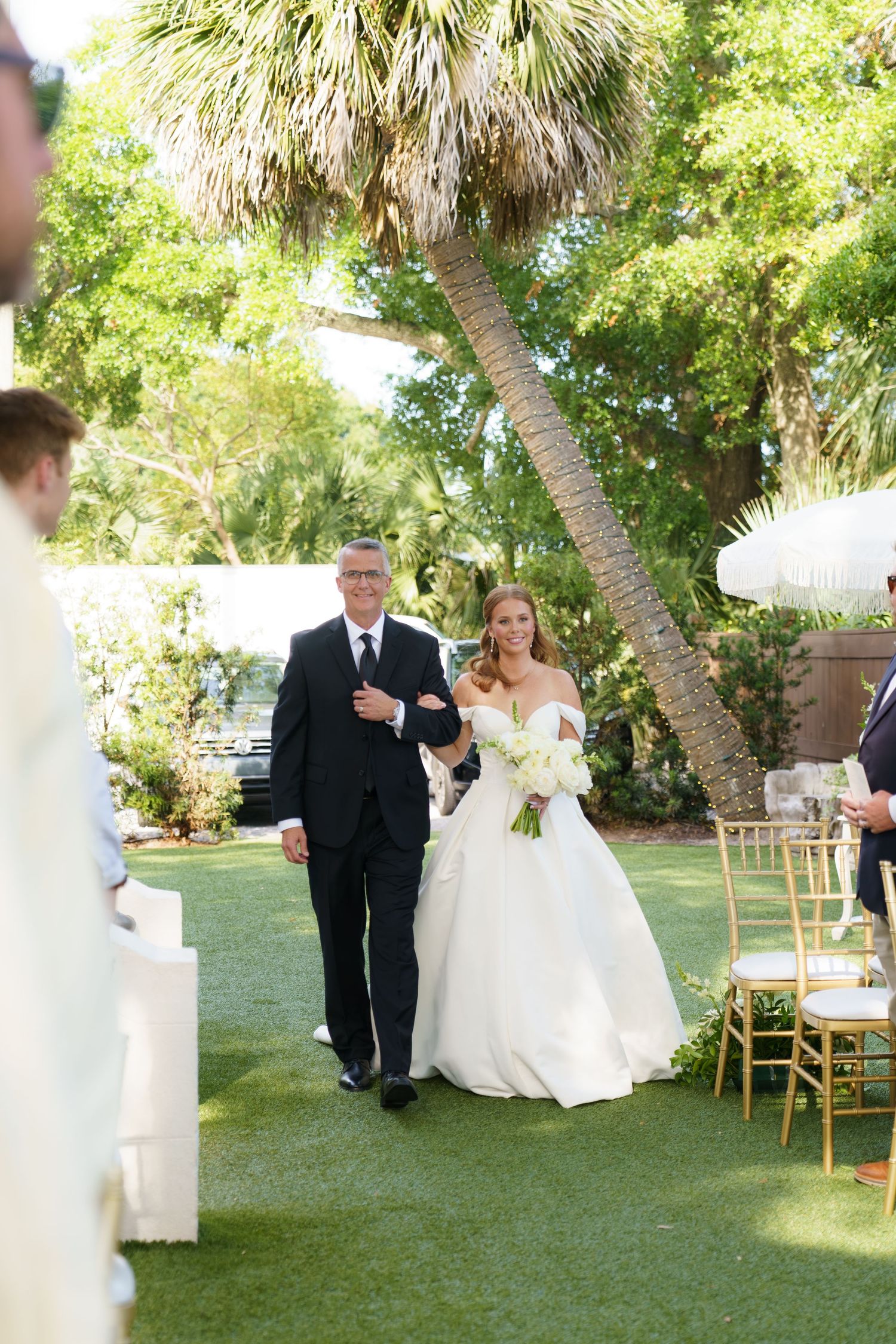 Bride in white off-shoulder gown walks down outdoor aisle with escort, surrounded by lush greenery and gold chairs.