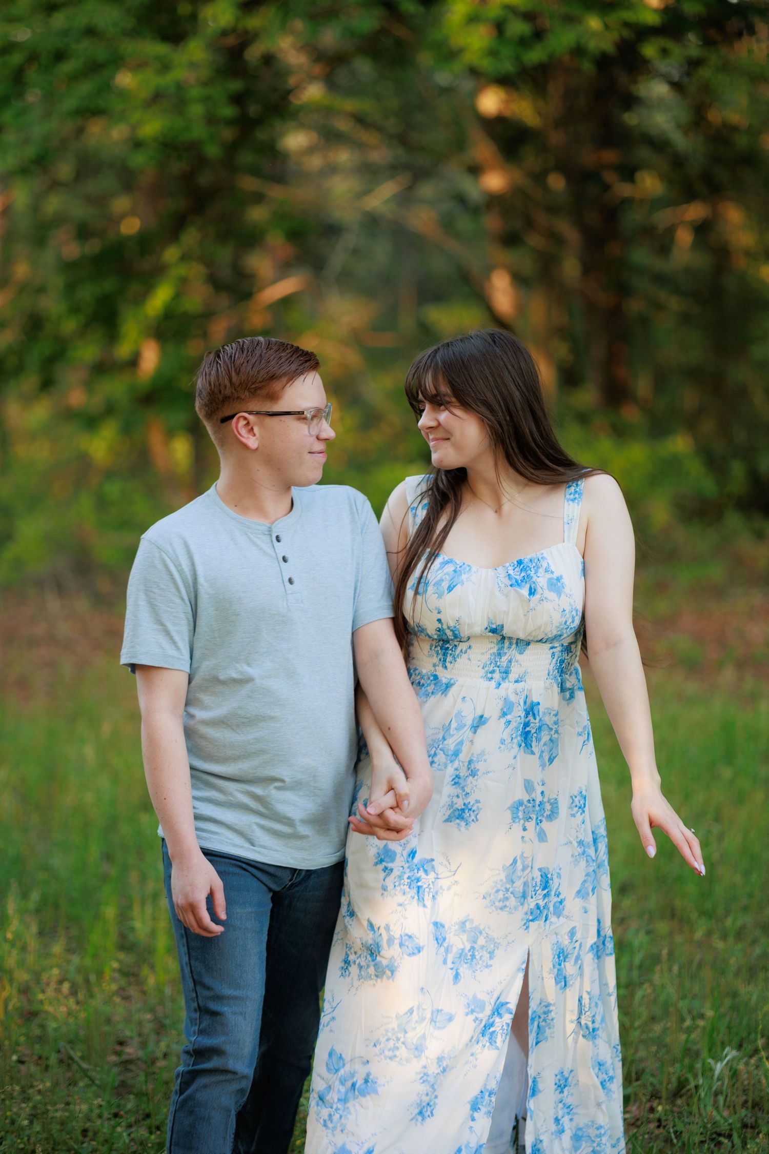 Couple holding hands and smiling at each other in a lush green park, woman in floral maxi dress, man in gray tee.