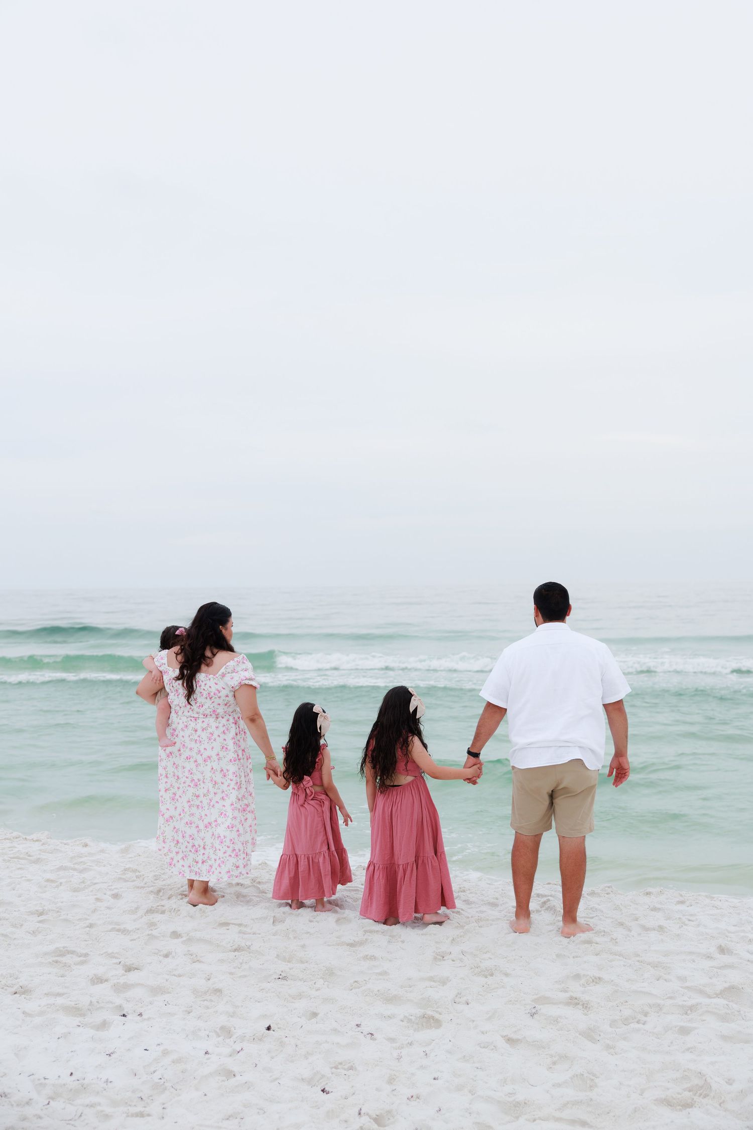 Family of five holding hands facing turquoise ocean waves on white sand beach, wearing coordinating pink and white outfits.