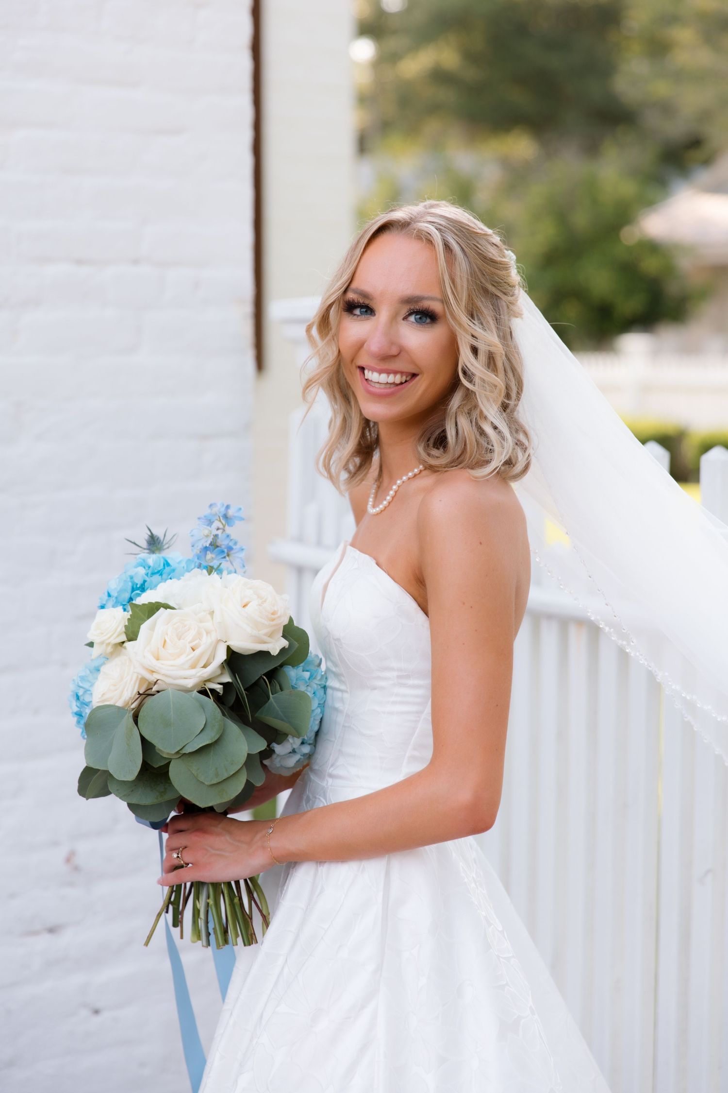 Bride in strapless wedding dress poses with blue and white bouquet against white railing.