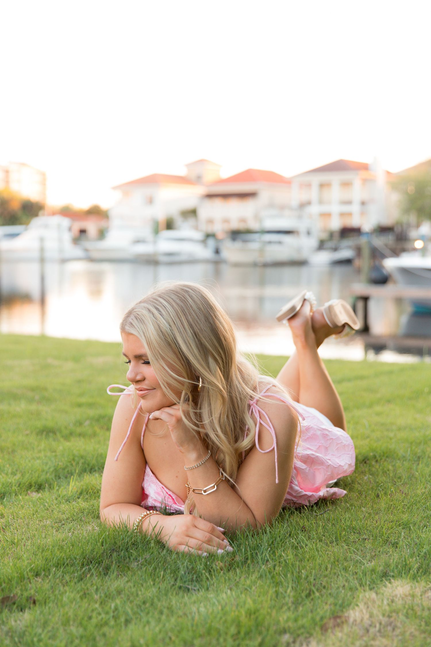 Woman in pink dress relaxes on grass near marina at sunset.